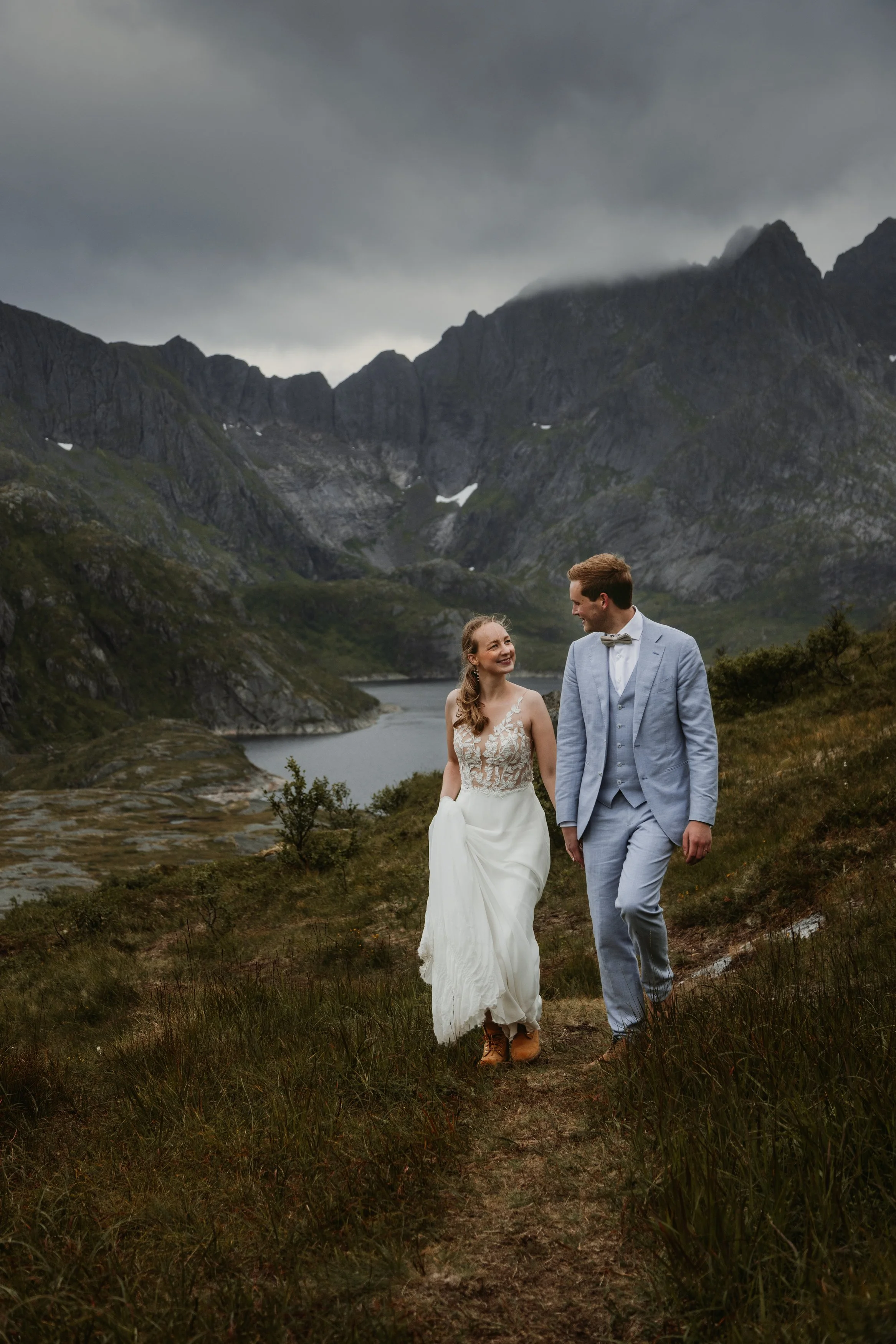 A bride and groom walking on a nature trail near a lake surrounded by mountains in Norway.