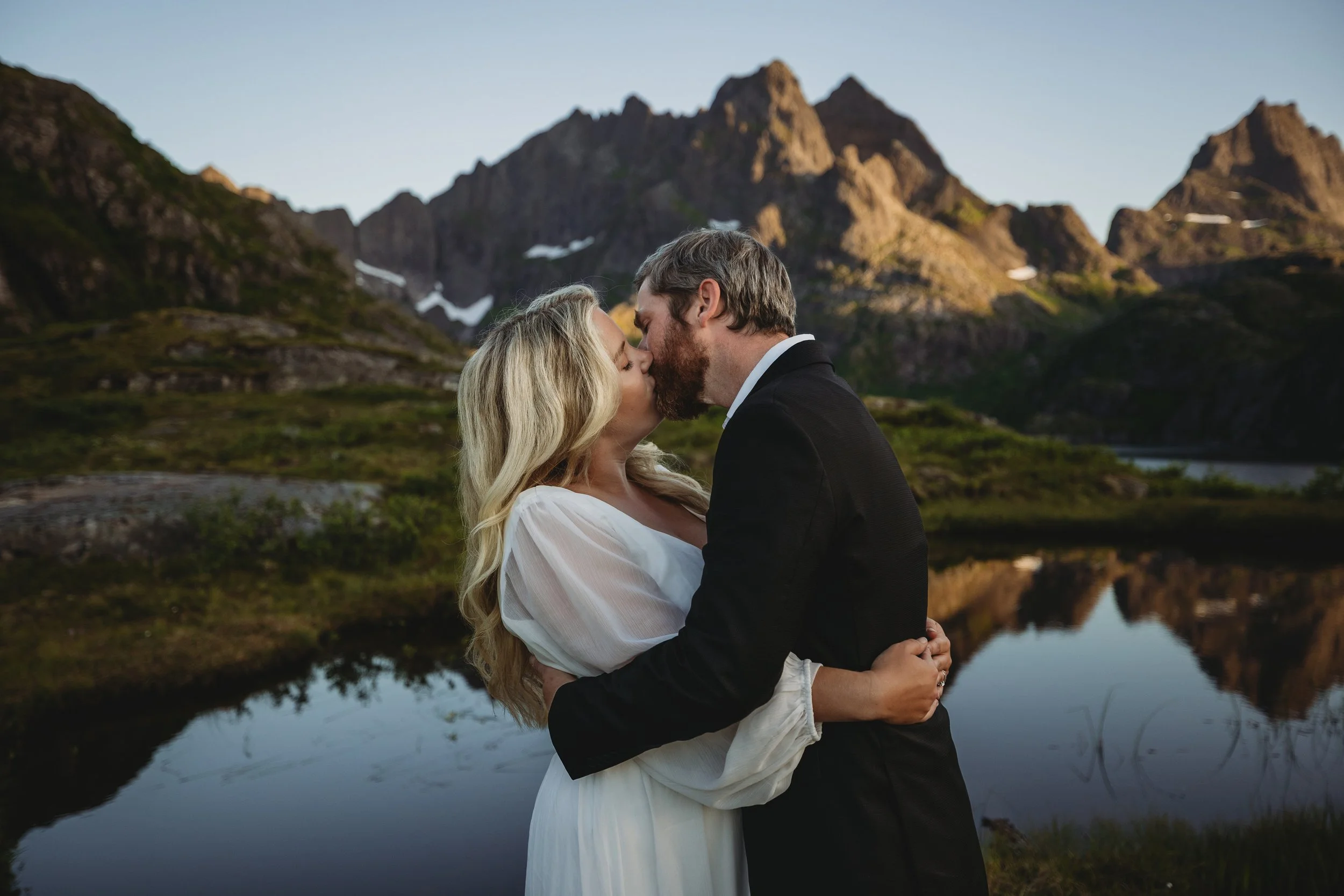 A couple kissing in front of a mountain landscape with a lake, mountains, greenery, and blue sky.
