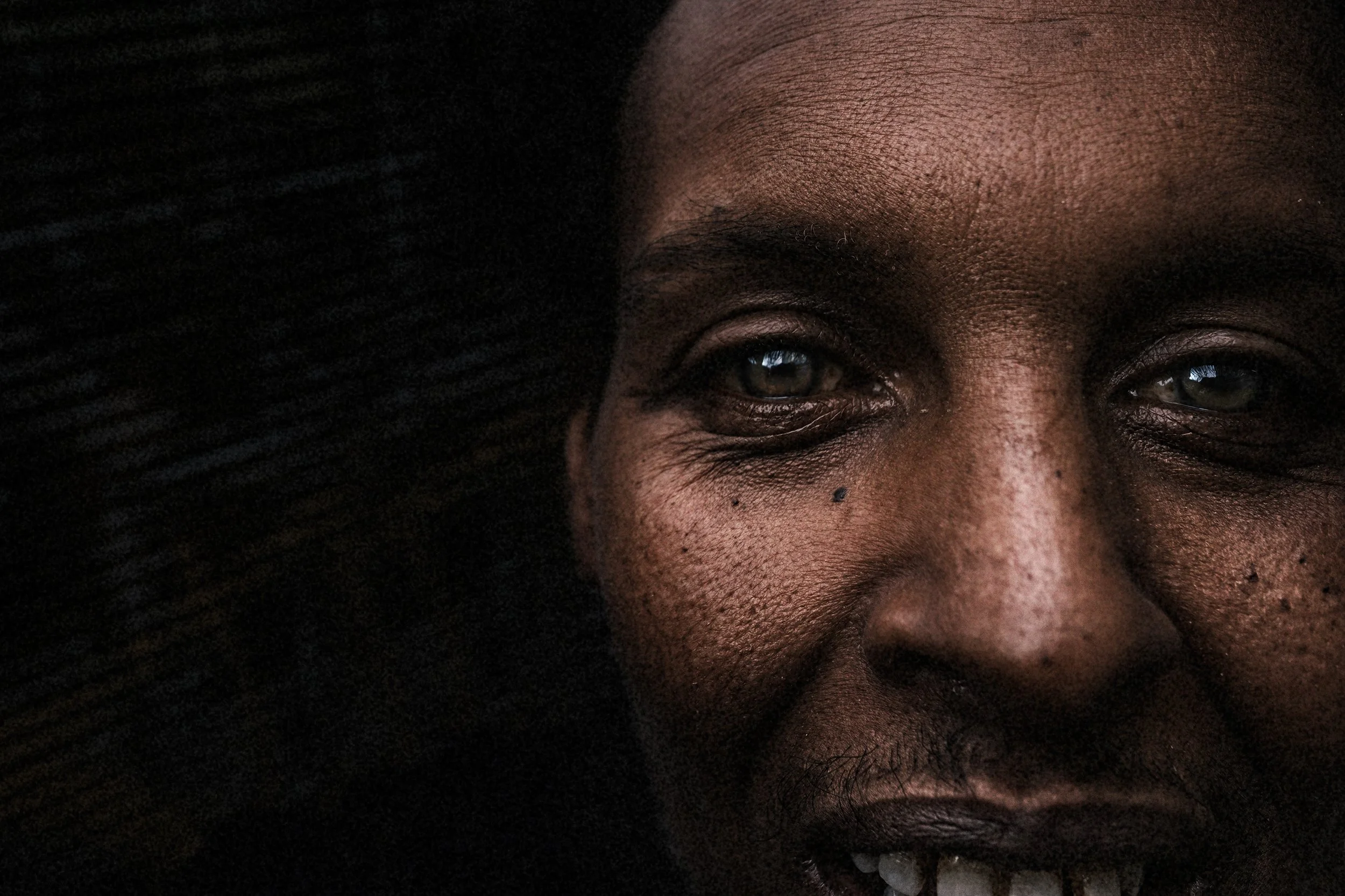 Close-up of a smiling person's face with dark skin, brown eyes, and natural hair, in low lighting.