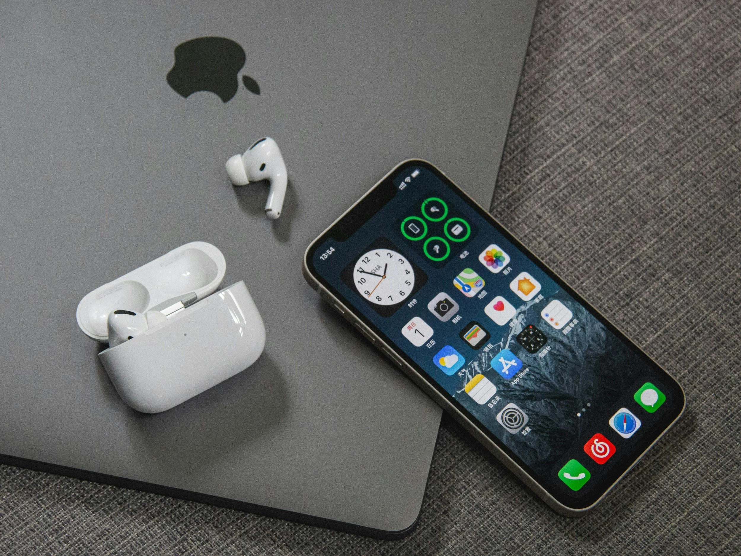 An iPhone with a black screen displaying apps, a pair of Apple AirPods in an open white charging case, all placed on a gray surface with a gray fabric background, and a gray Apple MacBook pro with an Apple logo.