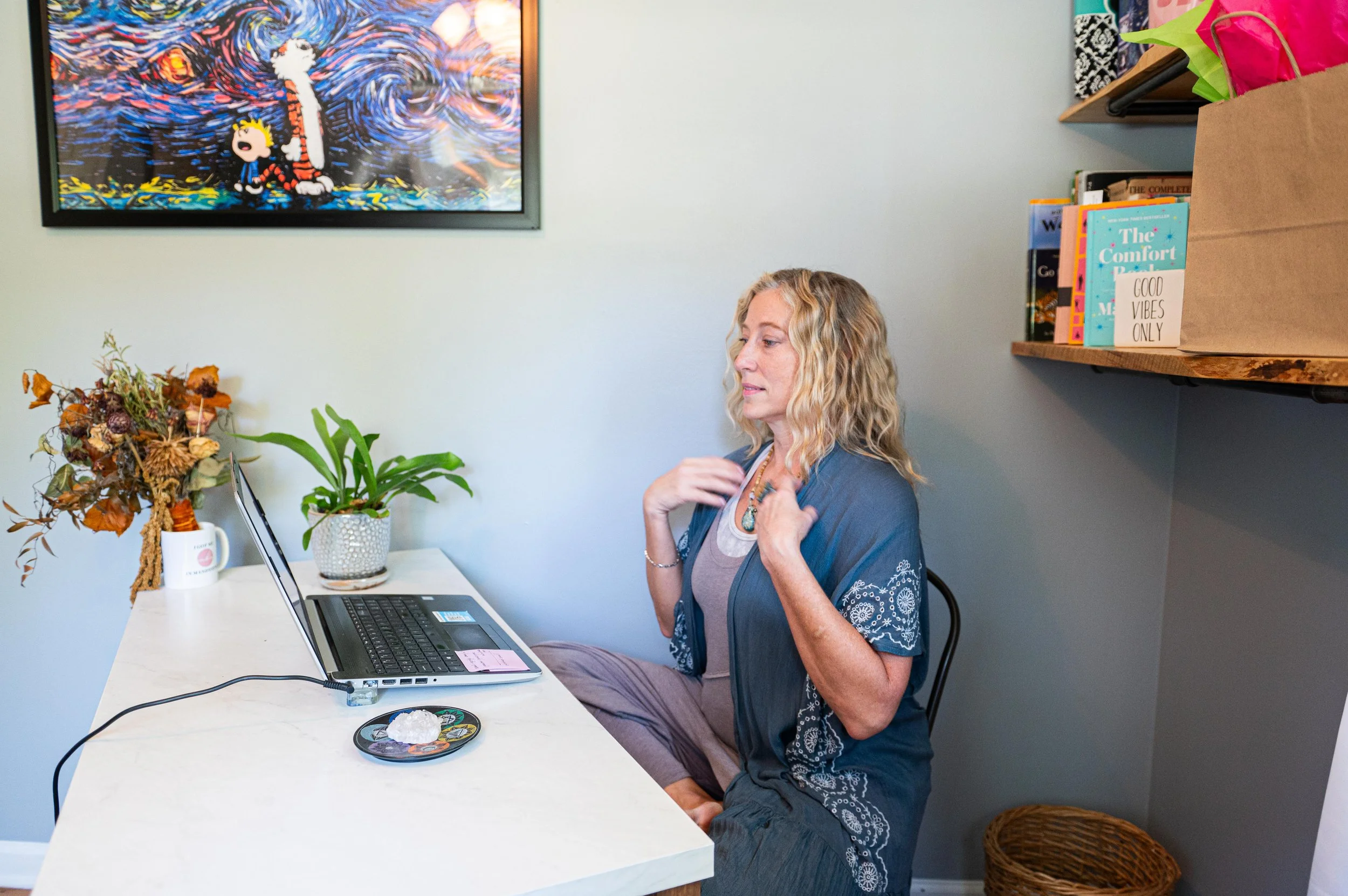 A woman with curly blonde hair sits on a chair at a white desk, adjusting her necklace. She is in a room decorated with plants, a colorful framed artwork of Calvin and Hobbes, and a shelf with books and a brown paper bag with colorful tissue paper.