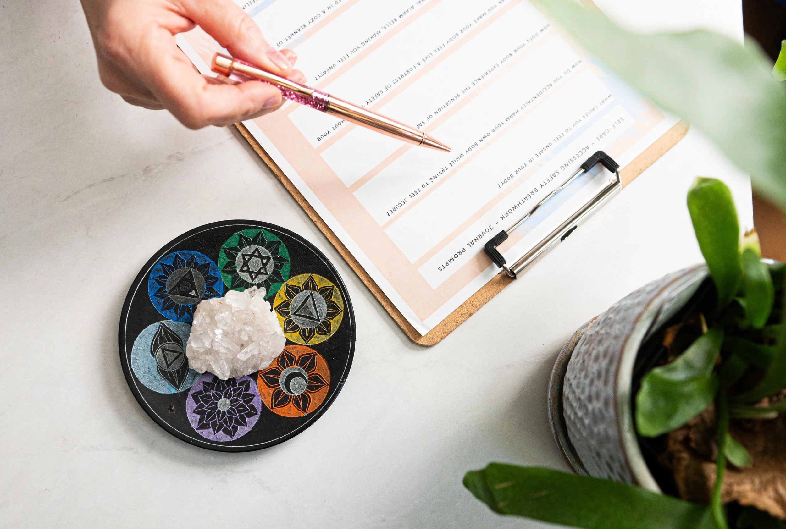 A person writes on a clipboard with a pink pen. On the desk, there is a black circular disc with colorful symbols and a white crystal. A potted green plant is also visible.