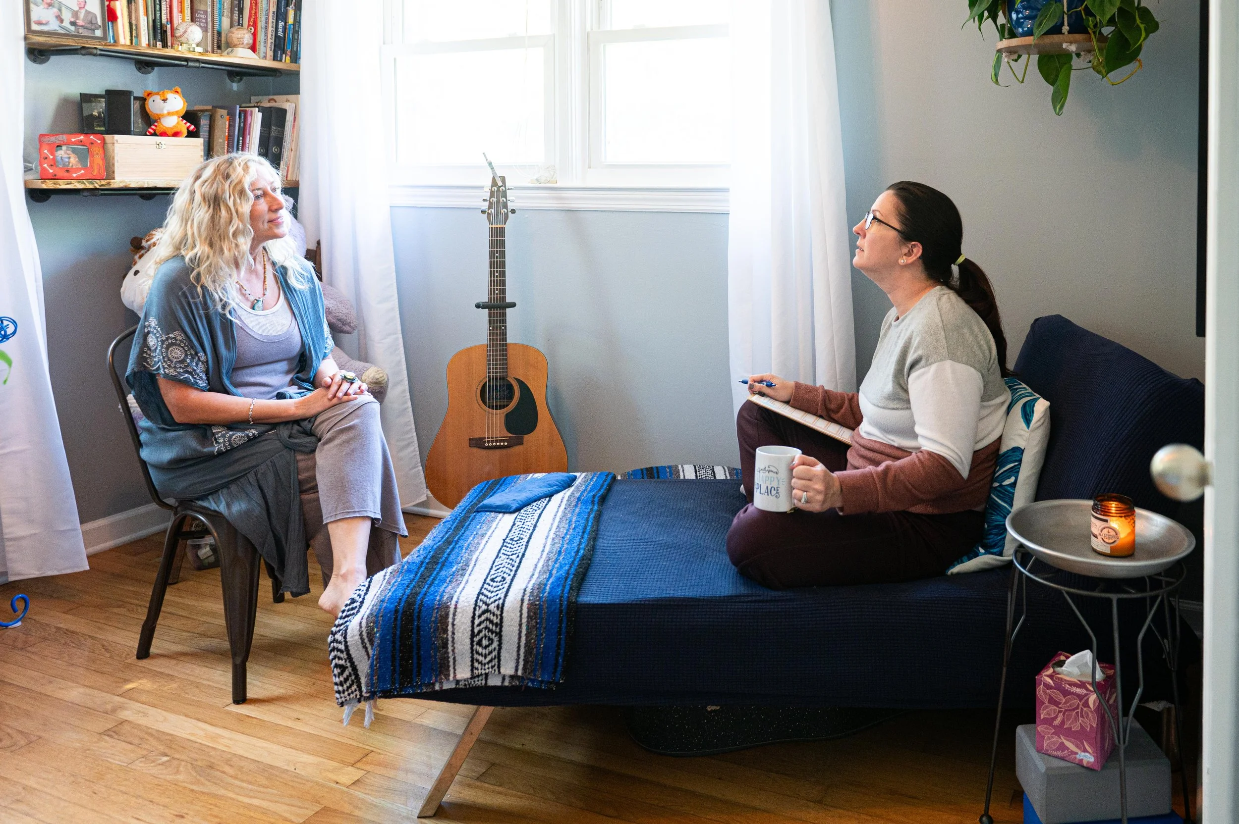 Two women having a conversation in a cozy room with a bed, bookshelf, guitar, and natural light from a window.