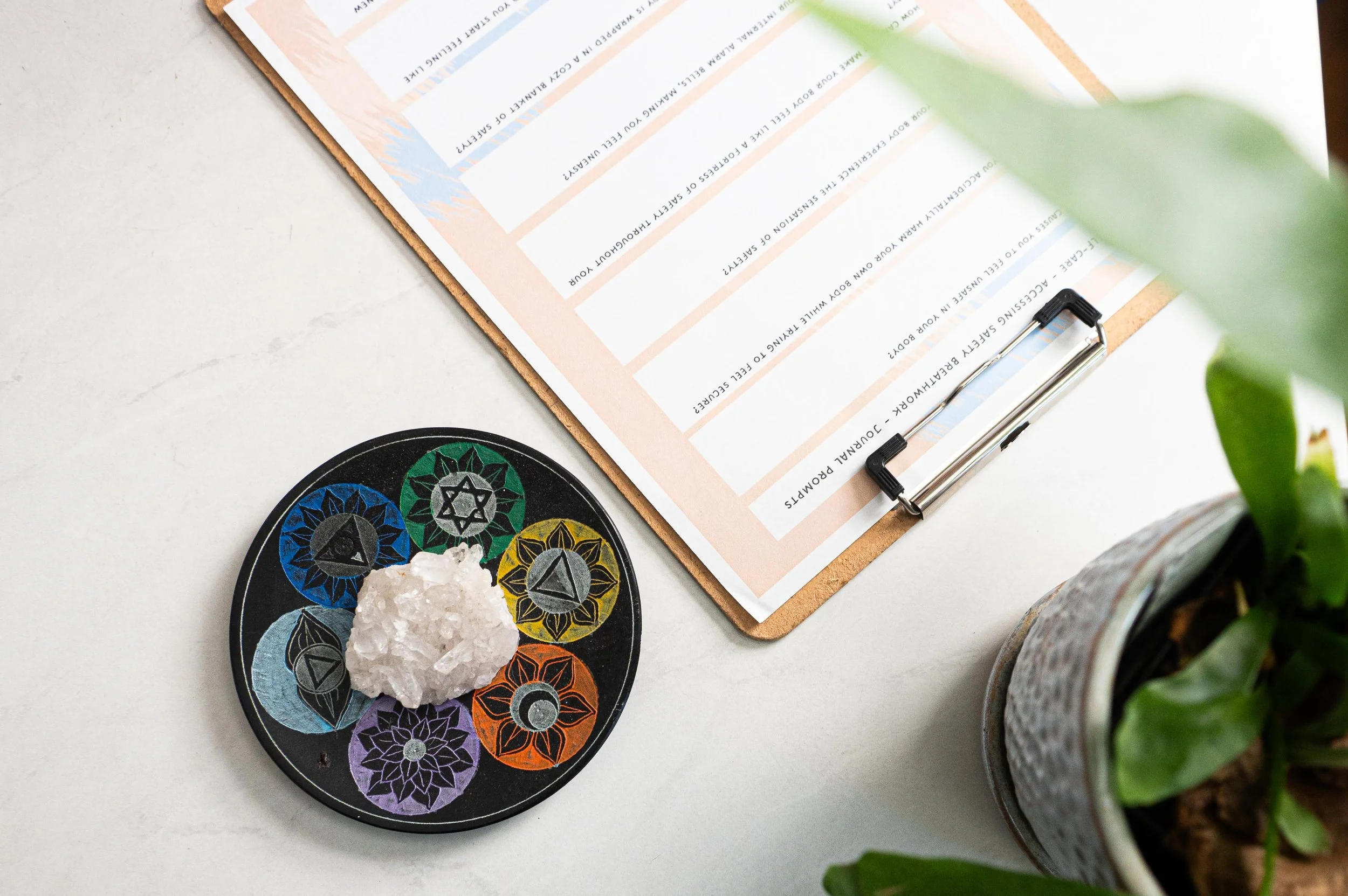 A black circular plate with colorful symbols and a white crystalline stone on top, coated with a botanical pattern of symbols in blue, green, yellow, orange, purple, and gray. The plate is next to a clipboard with a form on it, and a potted plant with broad green leaves partially visible on the right side.