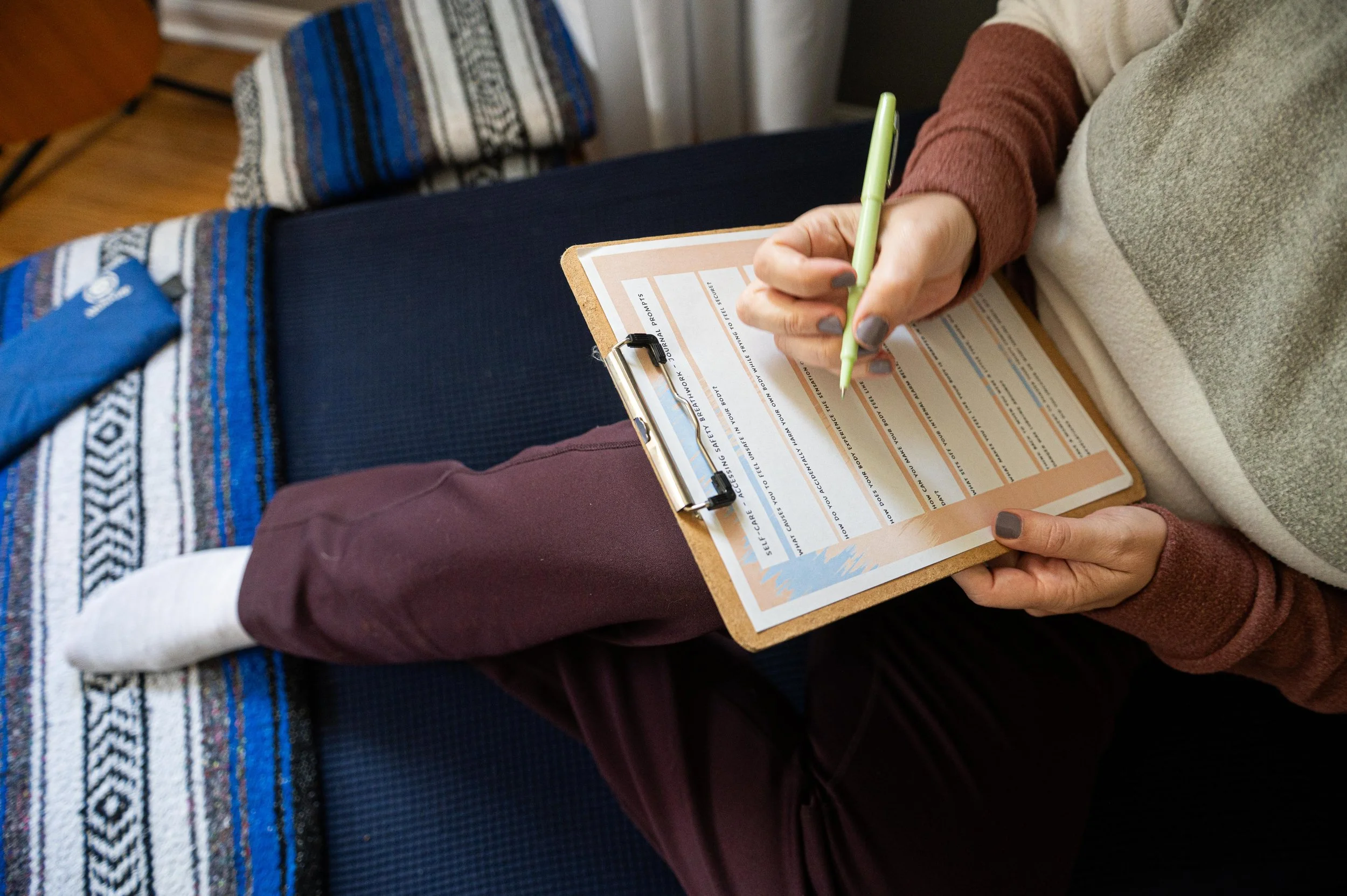 Person filling out a health concern or symptom checklist on a clipboard with a green pen, wearing maroon pants, a beige and brown sweater, sitting on a blue chair or bench.