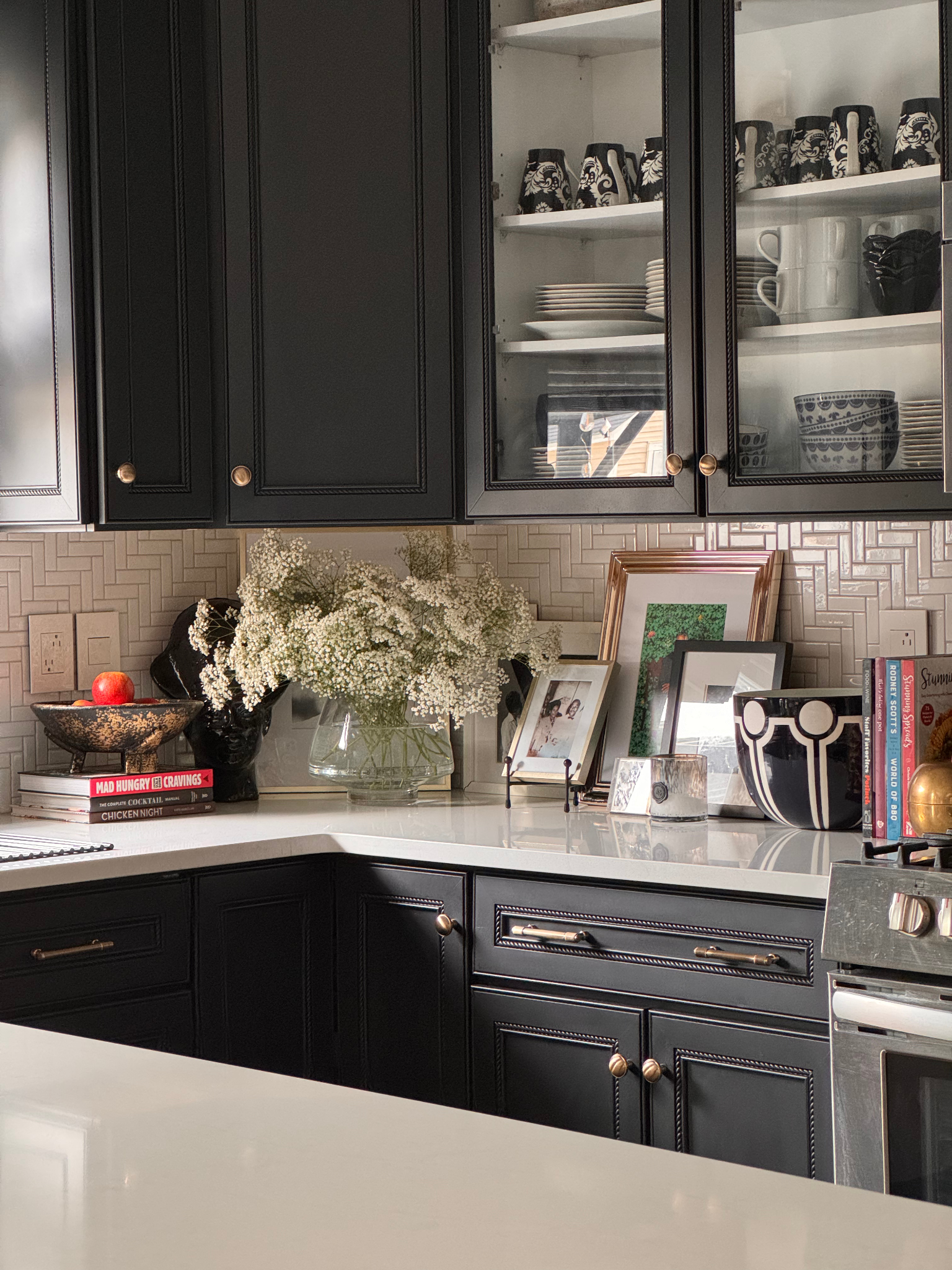 Kitchen with black cabinets, open glass-front upper cabinets displaying plates, bowls, cups, and mugs. A white countertop holds a vase of white flowers, framed photos, books, a bowl of apples, and various decorative items.