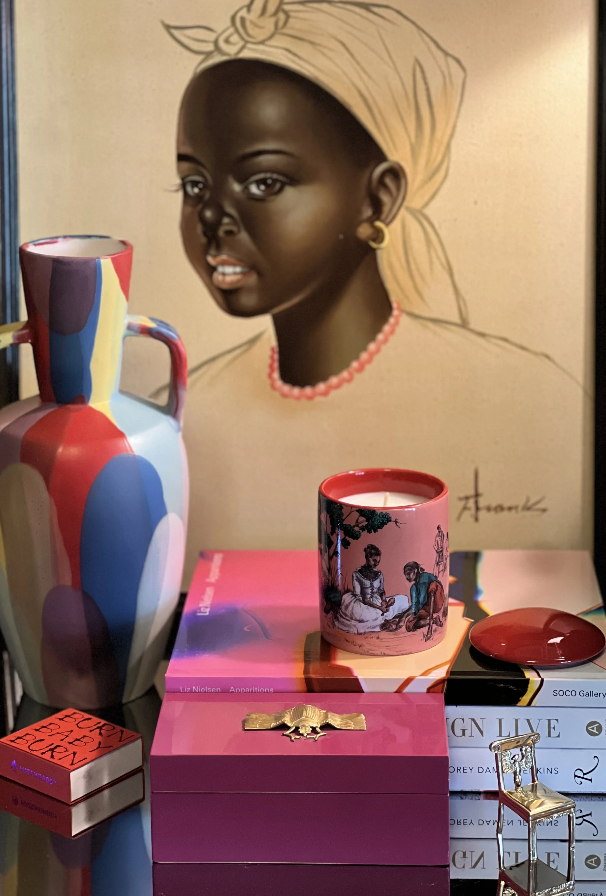 A colorful ceramic vase, a pink candle with an illustration, a small pink box with a gold decoration, a red dish, a stack of books, a miniature gold chair, and a large portrait of a woman wearing a headwrap and jewelry in the background.
