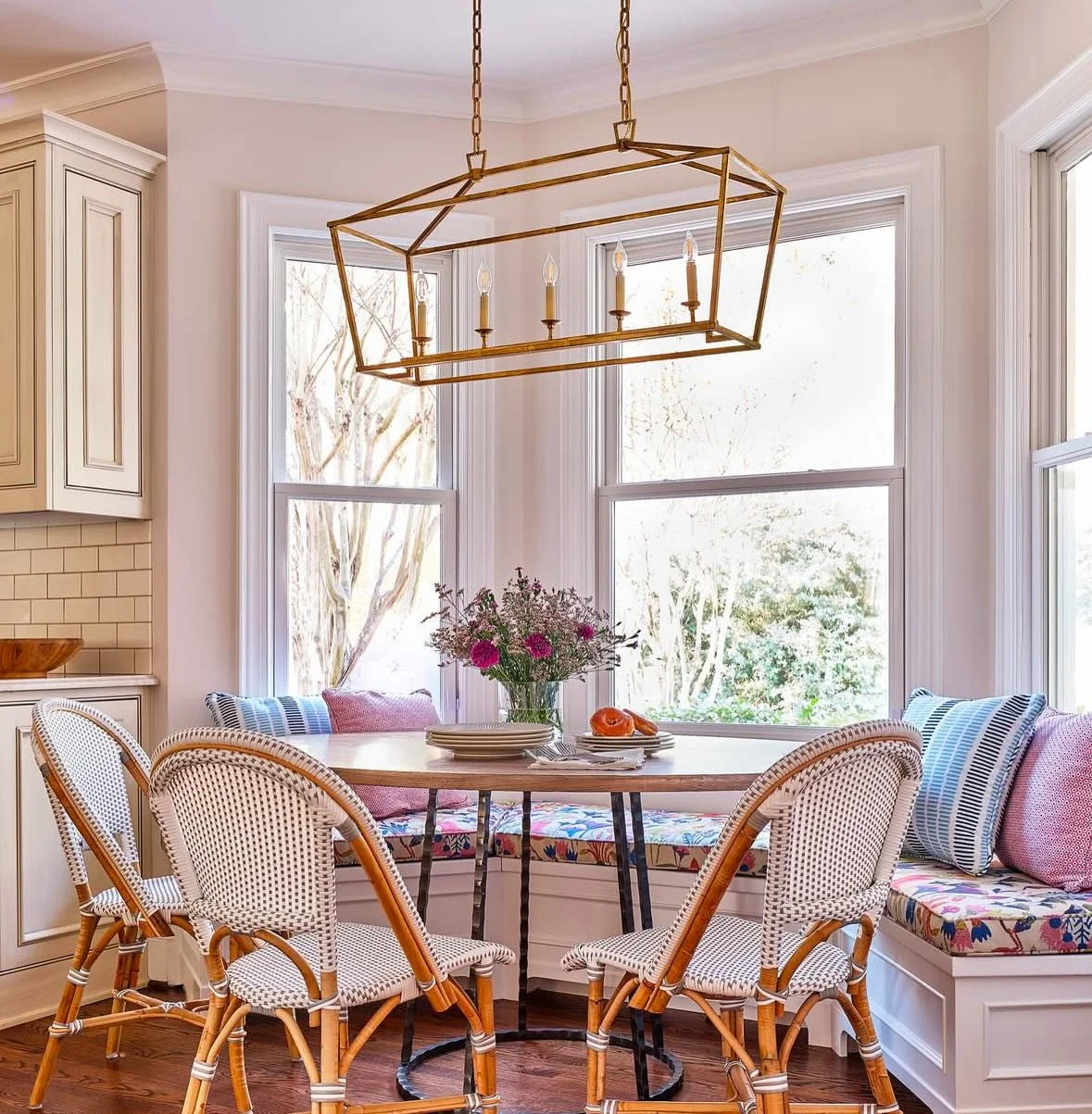 A bright dining area with a wooden table surrounded by rattan chairs and a built-in bench with colorful cushions, situated by large windows with a view of trees outside. A modern geometric chandelier hangs above the table, which is set with plates, a