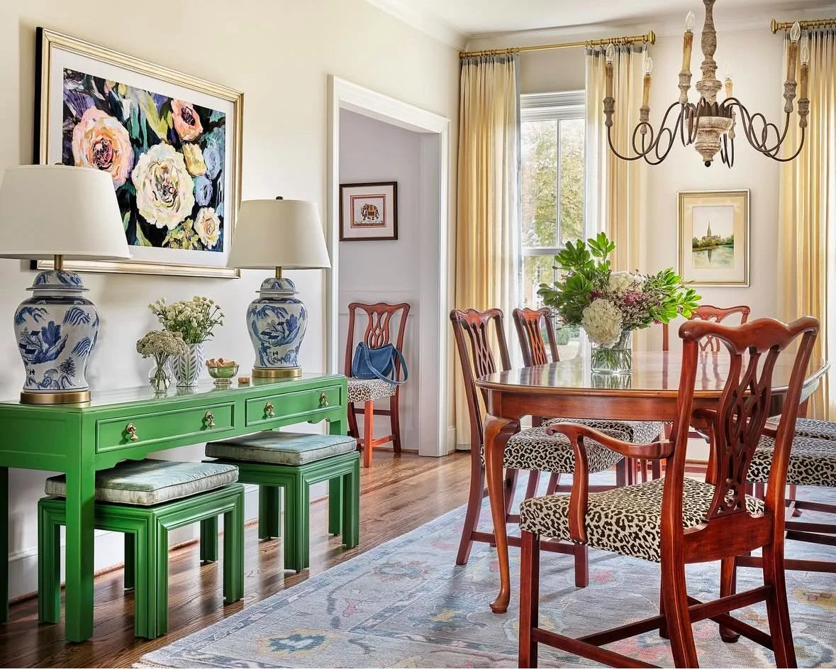 Living room with a green side table, two large blue and white lamps, floral artwork, a wooden dining table with chairs, floral centerpiece, curtains, chandelier, and windows.