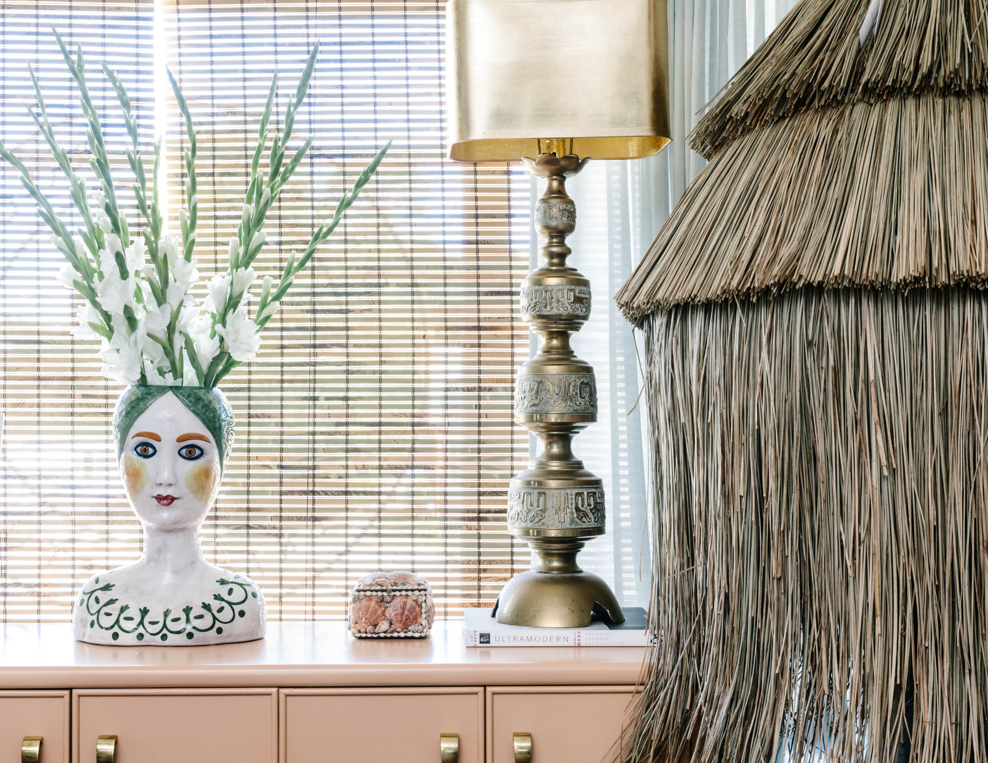Decorative interior featuring a vase with white flowers on a peach-colored sideboard, a small box, a tall ornate gold lamp, and a thatched structure partially visible on the right, with bamboo blinds in the background.