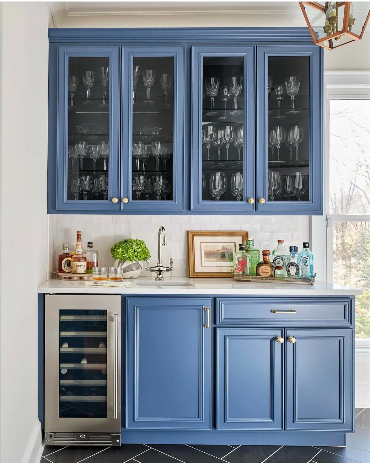 Blue kitchen cabinet with glassware, bar accessories, and a mini wine fridge beneath a white countertop near a window.
