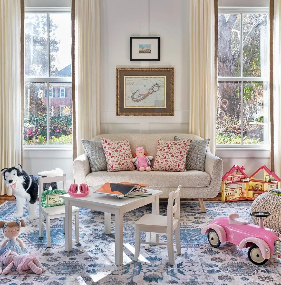 Bright living room with two large windows, white curtains, and a cozy beige sofa. The sofa has decorative pillows and a stuffed doll. In front of the sofa is a small white table with a book, pink headphones, and a green sign that says "Sweet Dreams."