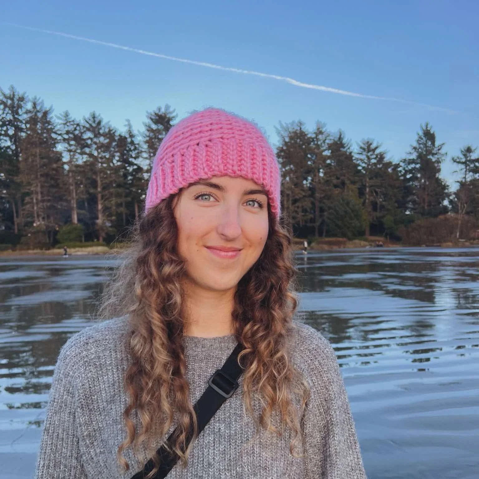 A close up photo of woman in pink toque and grey sweater on a beach.
