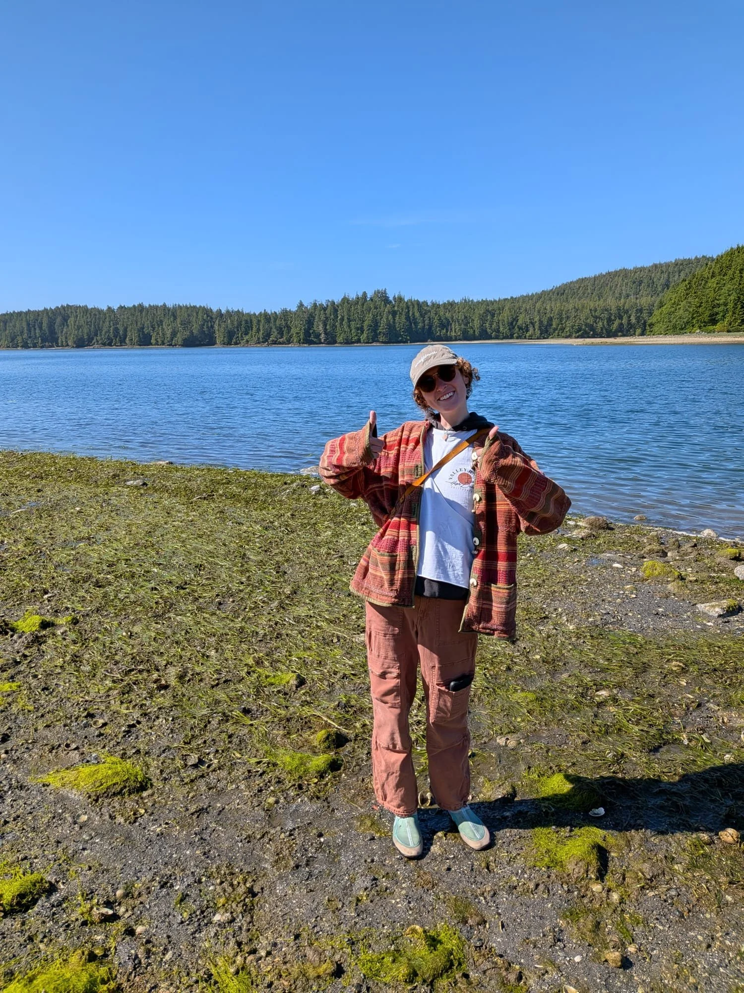 Person standing in front of a body of water making a thumbs up sign wearing a hat and sunglasses.