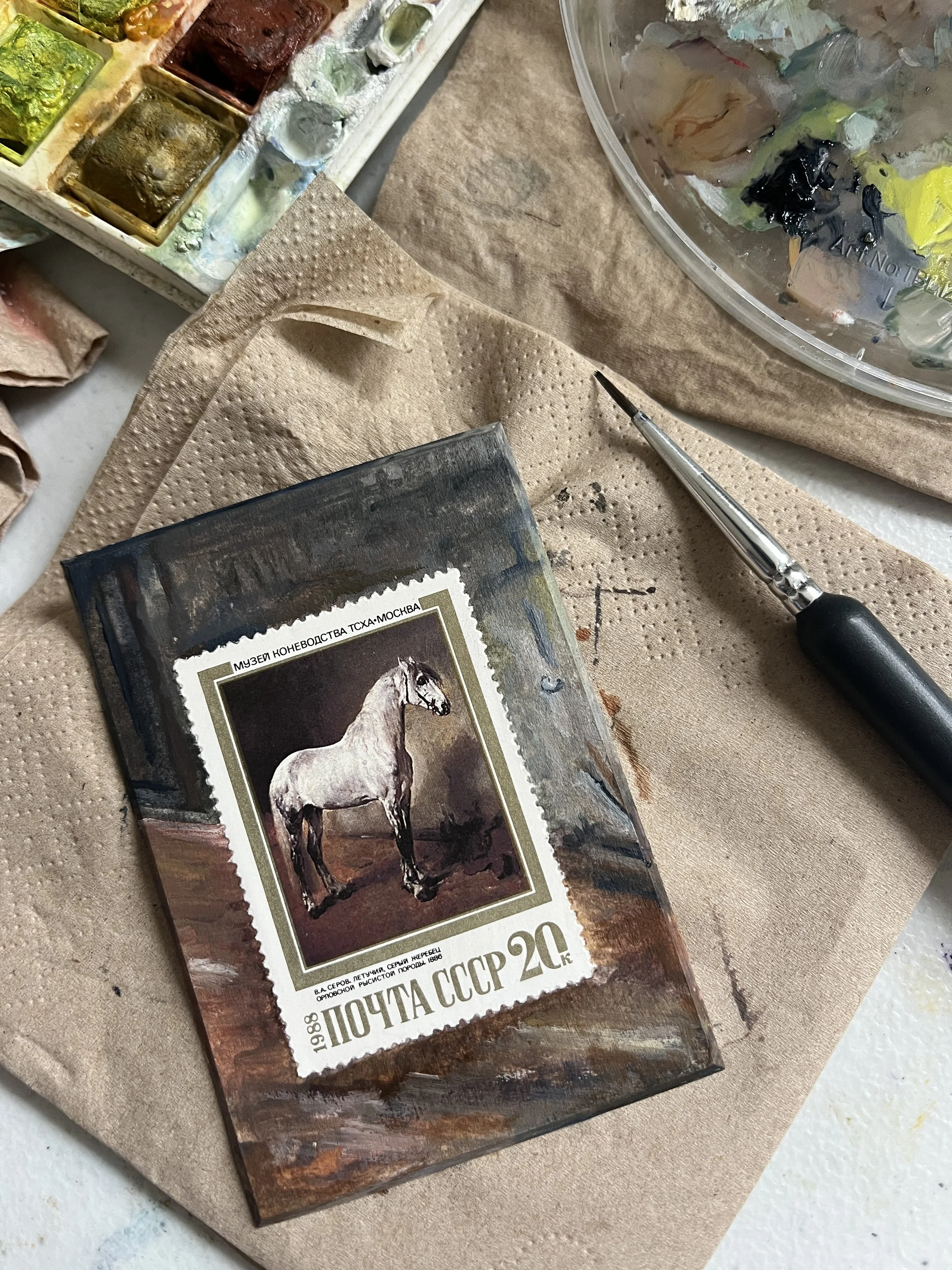 ussr horse painting on a postage stamp with a painted background showing a grey brown toned abstract painting of the stable background, paints and paintbrush are in view