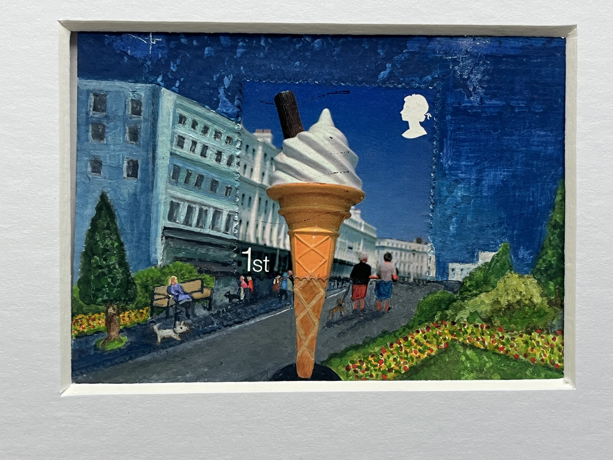 a large ice cream cone you would see outside and ice cream shop, with a flower garden in the foreground buildings in the back ground and people walking a dog in the road