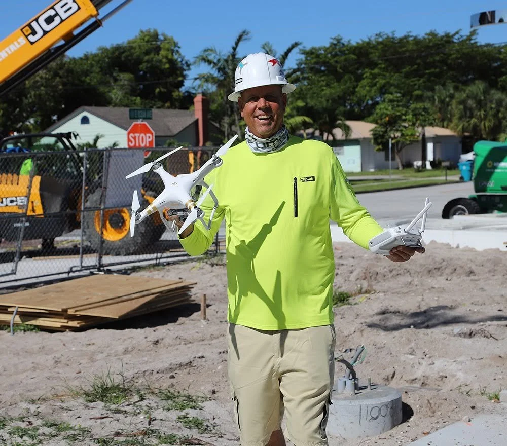 A smiling man in a yellow long-sleeve shirt, beige shorts, a white safety helmet, and a neck gaiter, standing outdoors at a construction site while holding a white drone and a remote control.