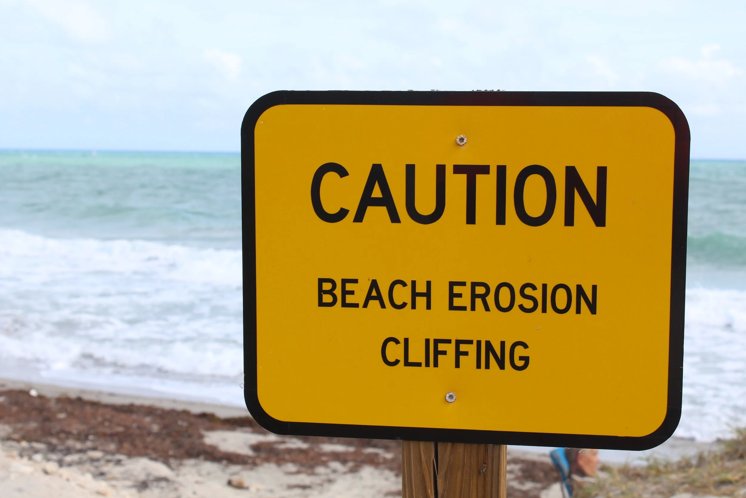 Yellow caution sign warning about beach erosion and climbing cliffs, with ocean and cloudy sky in the background.