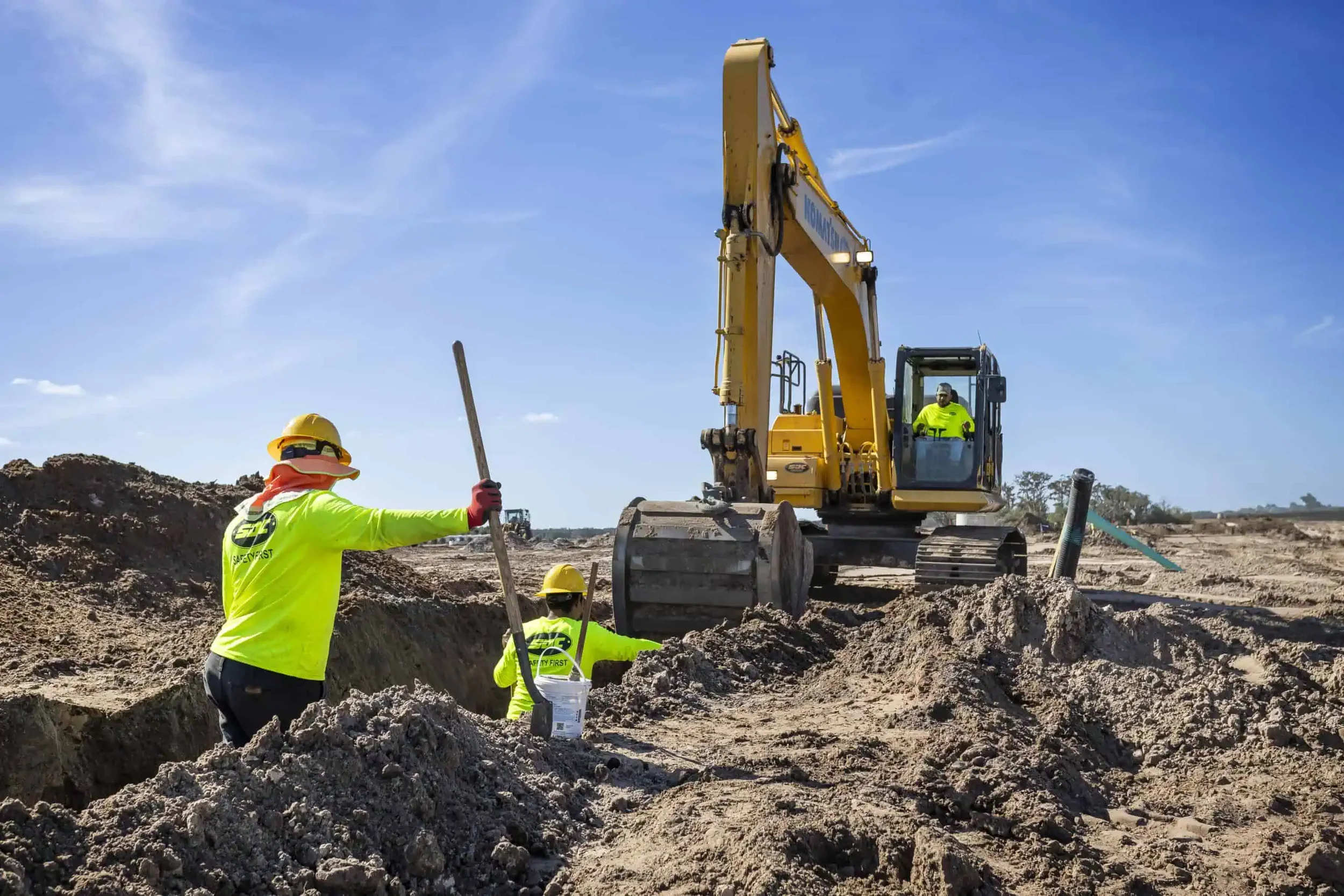 Construction workers in bright yellow safety gear working on a site with an excavator under a blue sky.