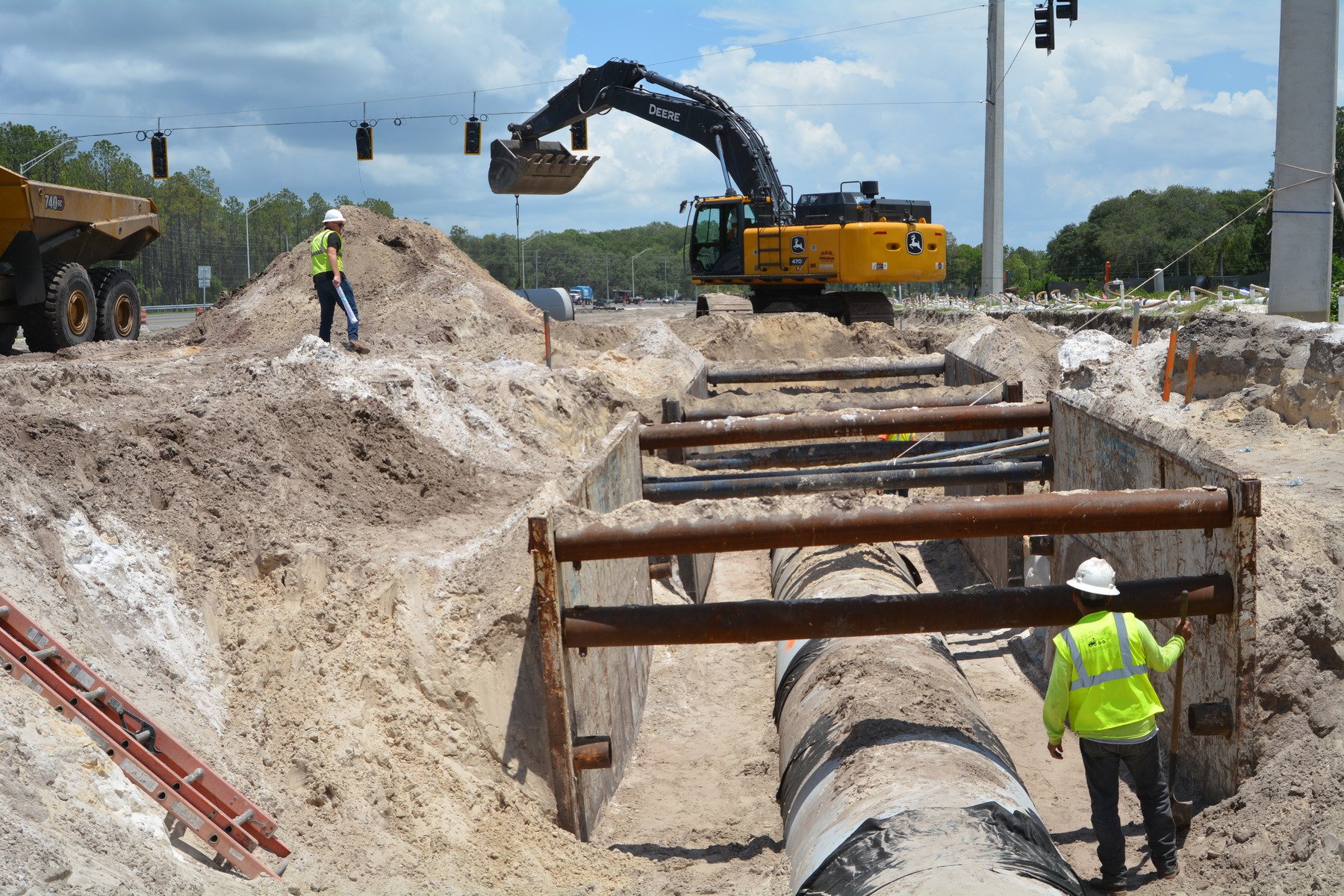 Construction workers and equipment working on underground pipeline installation on a road, with excavated dirt, a yellow excavator, and large pipes.