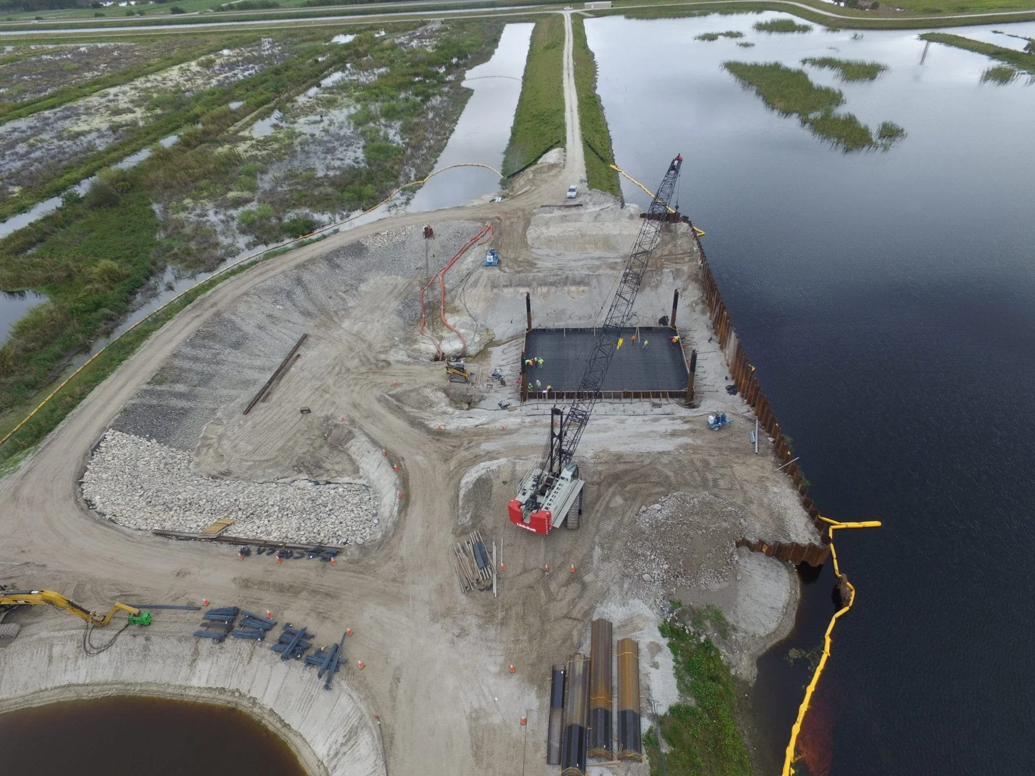 Construction site near a body of water with cranes, heavy machinery, and dirt roads under development.