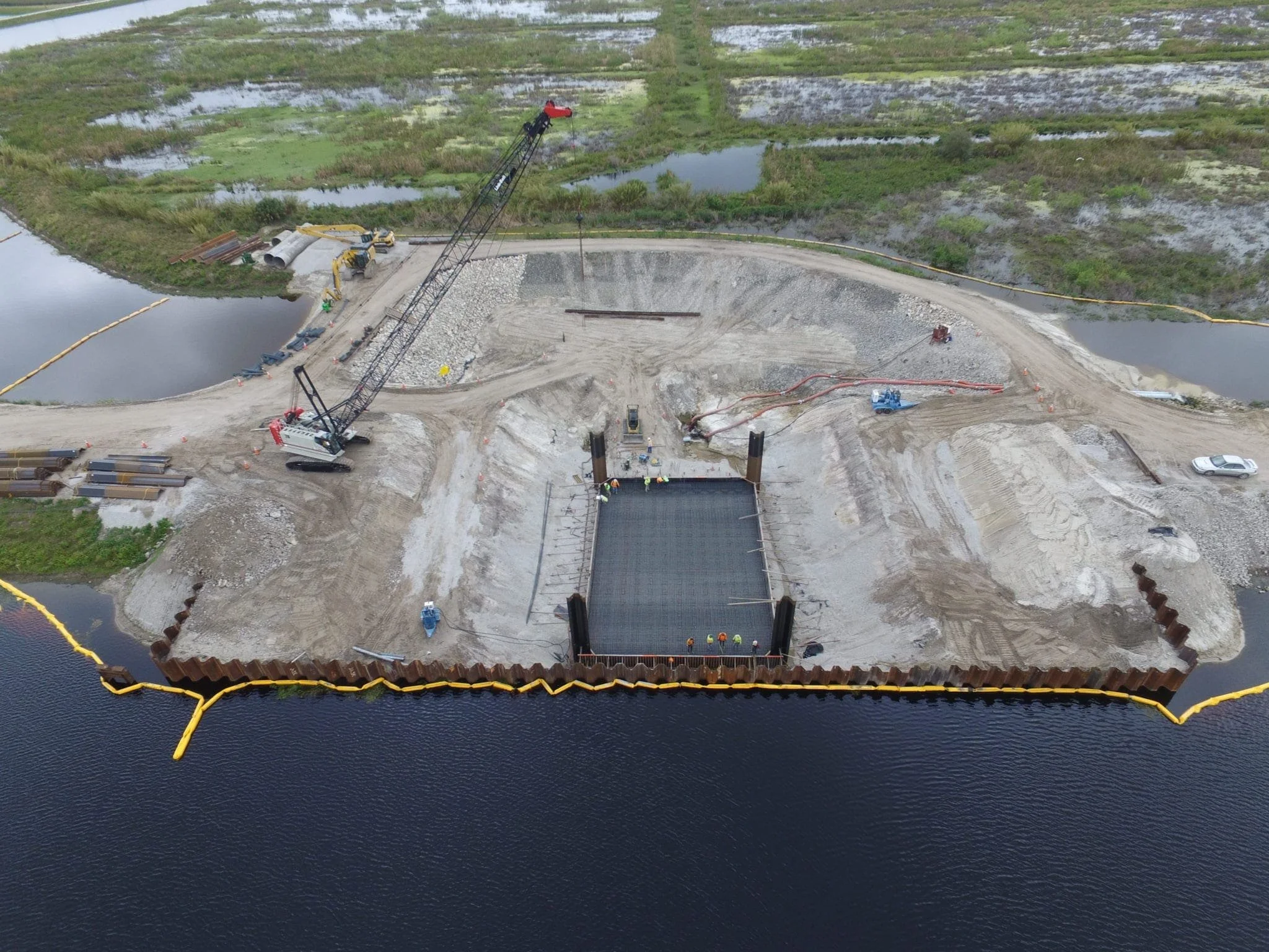 Aerial view of a construction site on water, with a large crane, construction workers, and a perimeter barrier, surrounded by wetlands and water bodies.