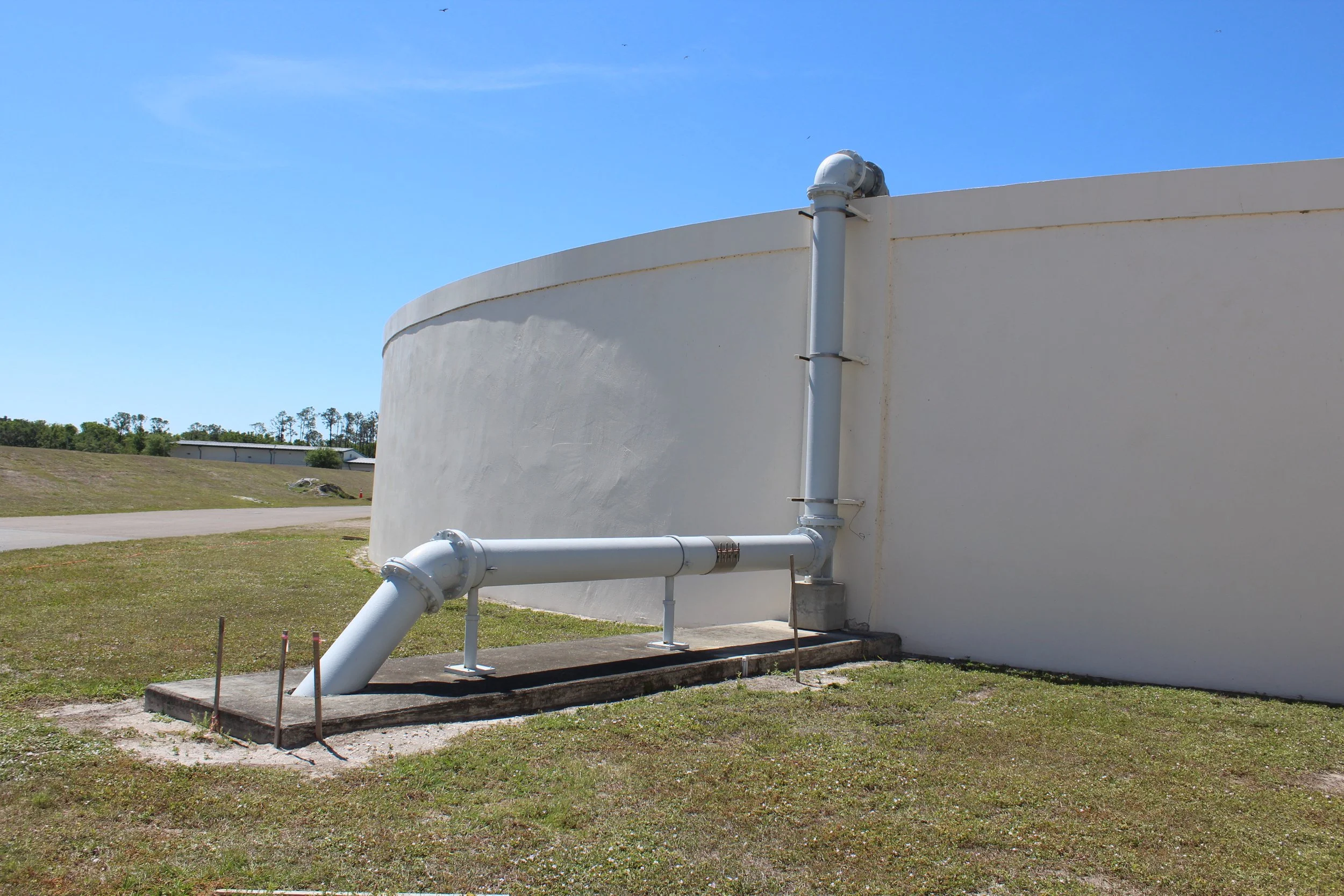 A large, white, cylindrical water storage tank with an attached white pipe system outdoors on a sunny day with a blue sky and some trees and buildings in the background.