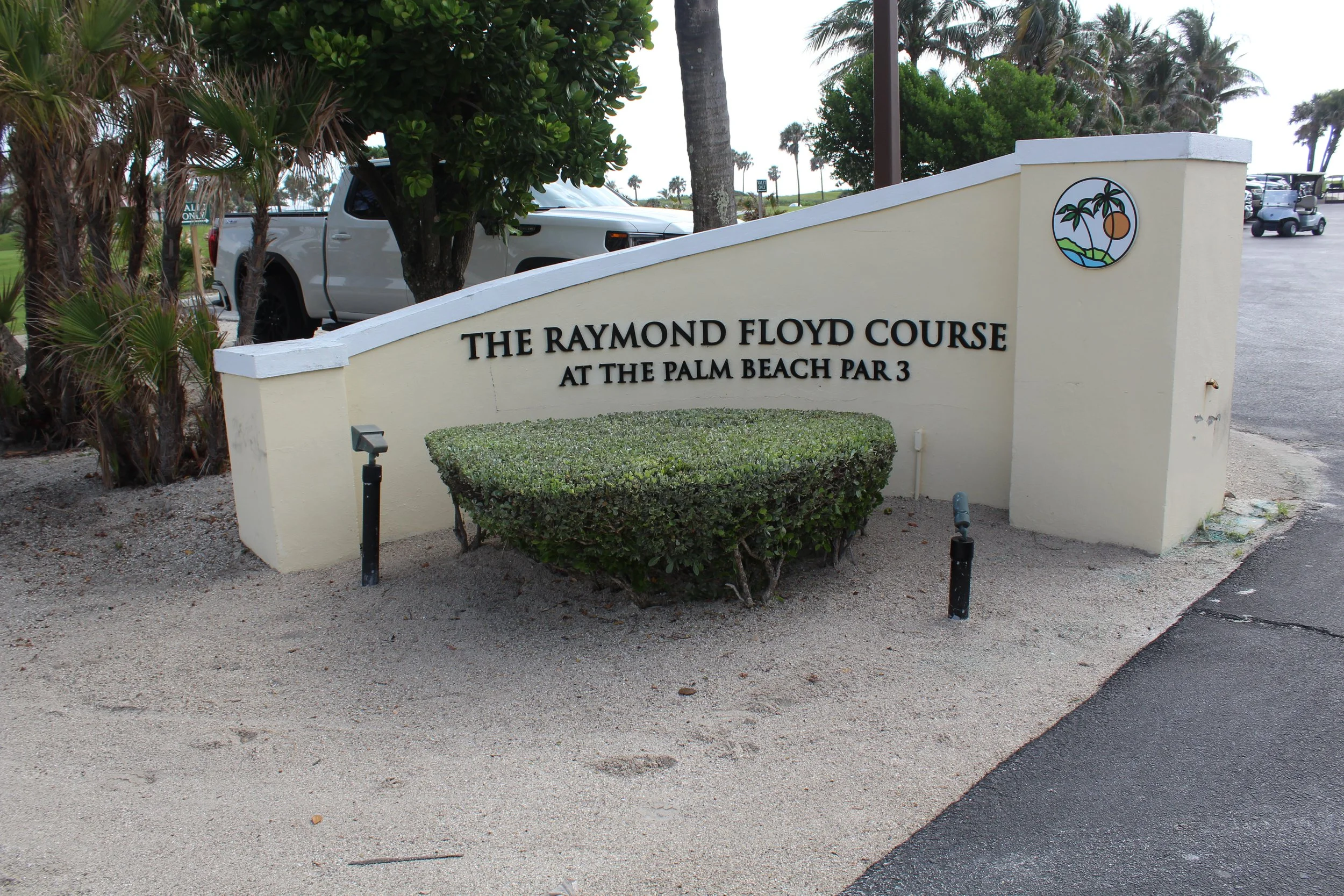 Sign for The Raymond Floyd Course at The Palm Beach, featuring a circular logo with palm trees, a sun, ocean waves, and golf course landscape.