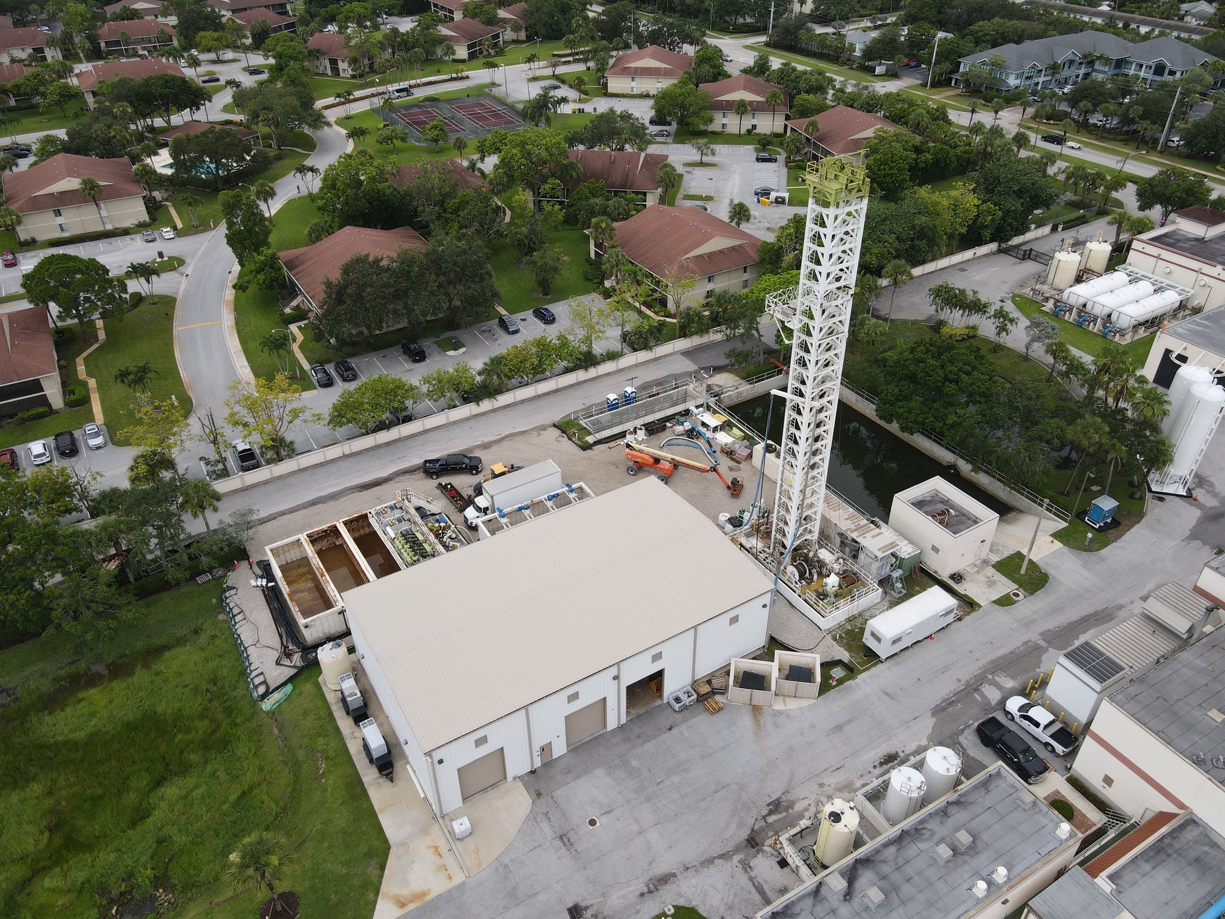 Aerial view of a residential area with multiple houses, parking lots, tennis courts, and lush green trees, alongside an industrial or commercial building with storage tanks.