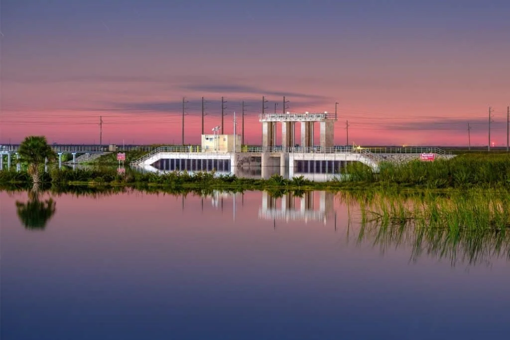 A power substation by a body of water at sunset with pink and purple sky.
