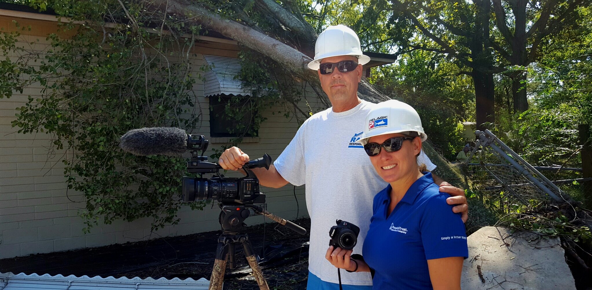 Two people in hard hats and sunglasses standing near a fallen tree that has broken a utility pole, which is leaning against a house, with landscaping and trees in the background.