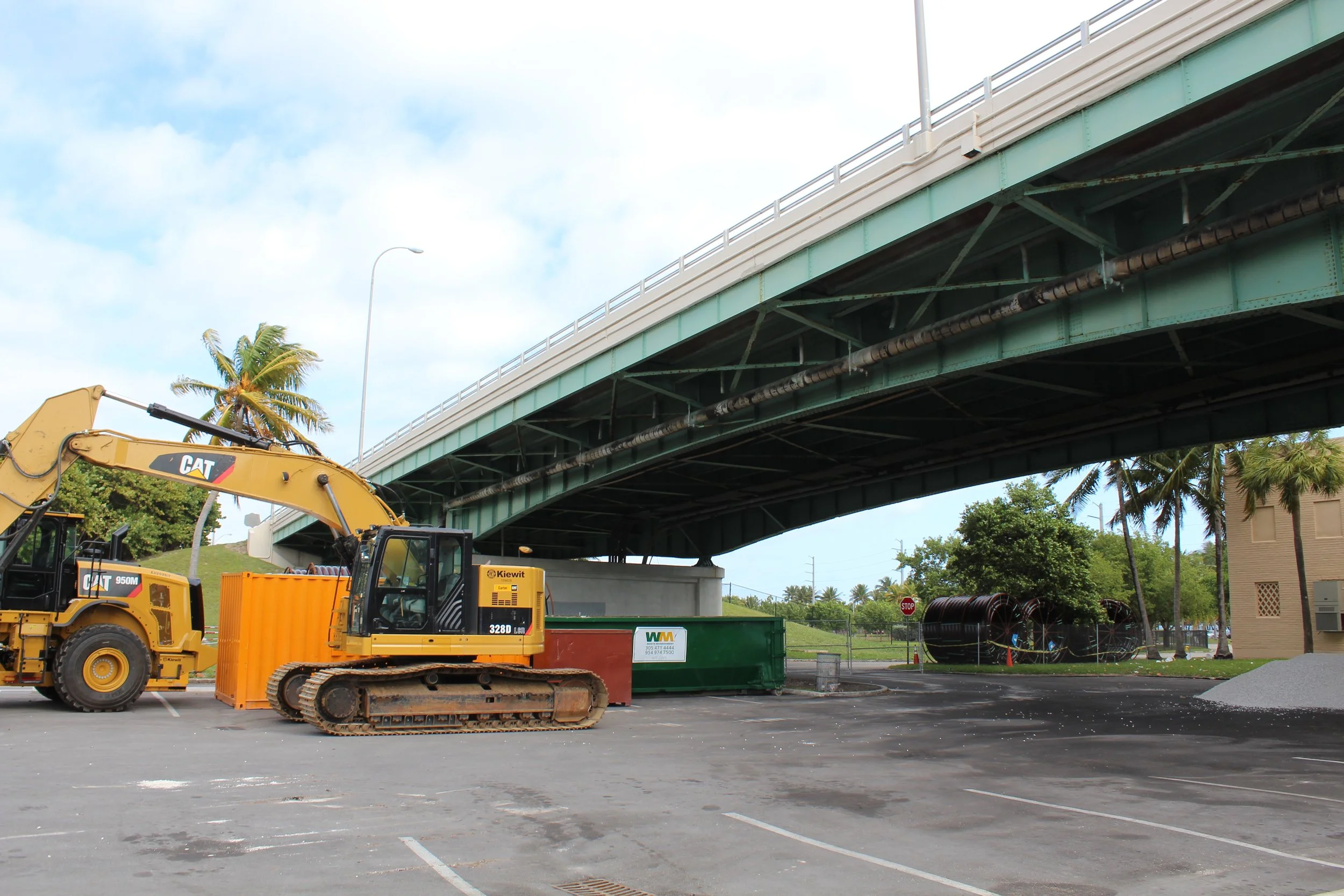 Construction site under a bridge with machinery, including a yellow Caterpillar excavator and an orange dumpster, in a parking lot with palm trees in the background.