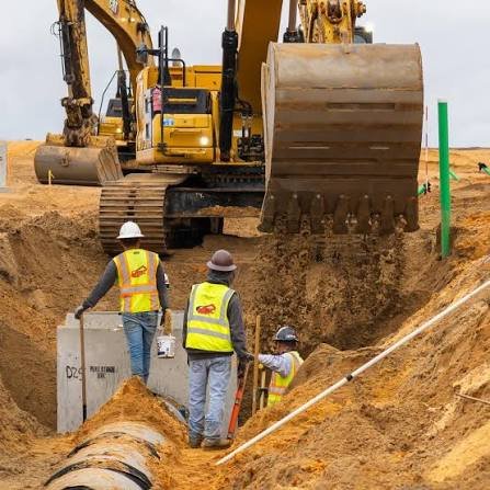 Construction workers in safety vests and helmets working on underground utility installation with excavator digging the ground.