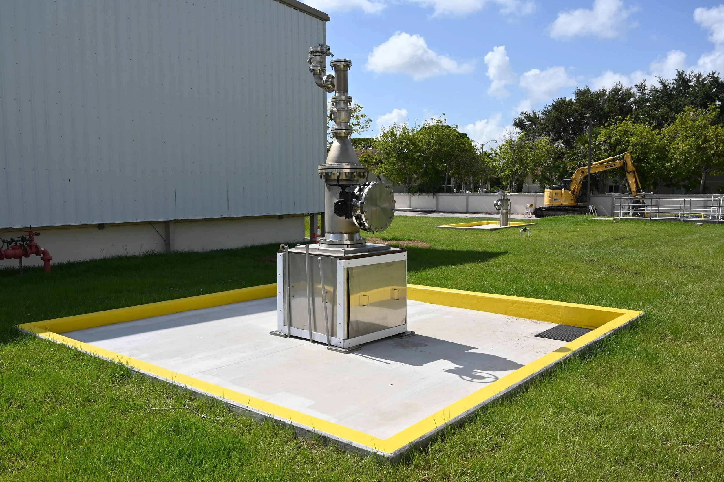 Industrial equipment with pipes and valves outside a building, surrounded by grass and trees, with a yellow safety barrier on a concrete slab.