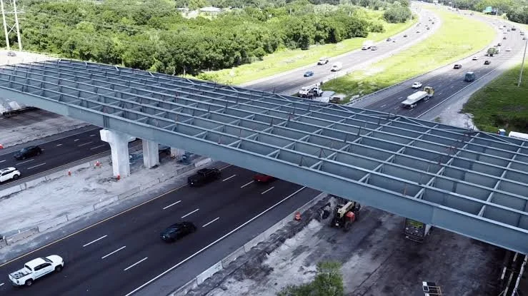 Construction of an overpass or bridge above a highway with visible support columns, and multiple lanes of cars and trucks on the road below, with trees and greenery in the background.