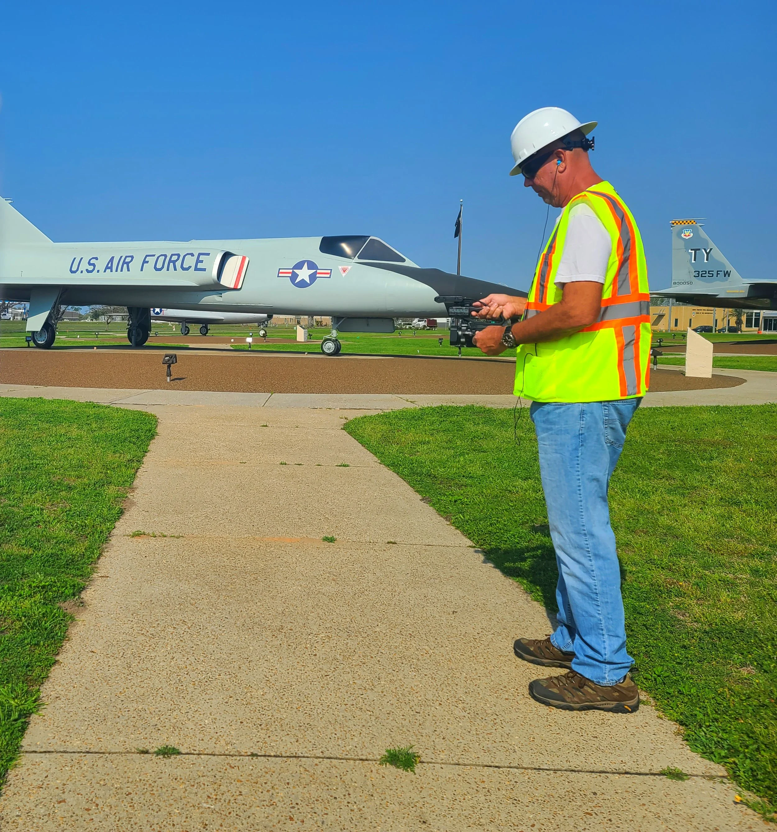 A man in a white hard hat, safety vest, and blue jeans is standing on a sidewalk, controlling a remote drone. Behind him is a U.S. Air Force fighter jet with the markings "U.S. AIR FORCE" and the tail number "325 FW." The scene is outdoors on a brigh