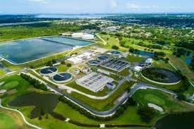 Aerial view of a wastewater treatment plant with multiple tanks, clarifiers, and green fields around it, surrounded by water and trees.
