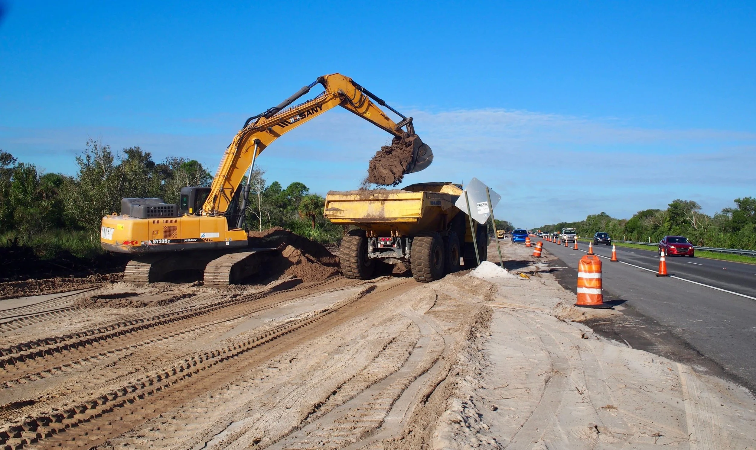 Construction site on the side of a highway with an excavator loading dirt into a dump truck, orange traffic cones, and cars driving on the road under a blue sky.