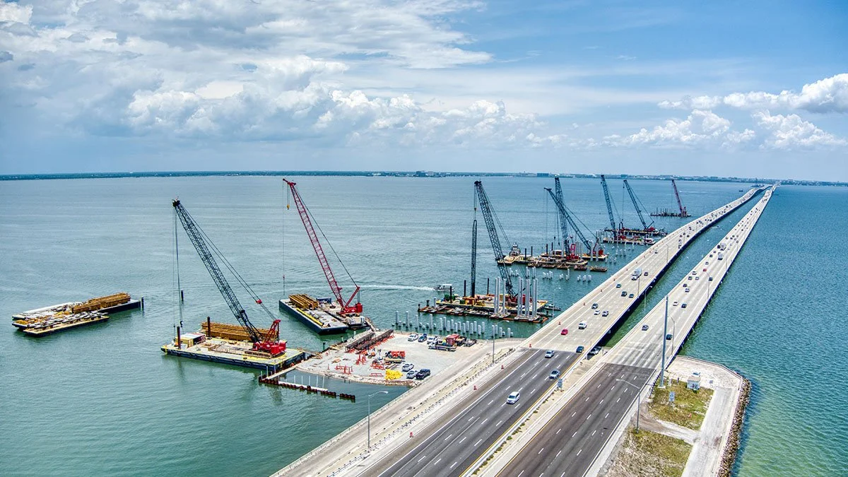 A large bridge under construction over a body of water, with cranes and construction equipment on floating platforms.