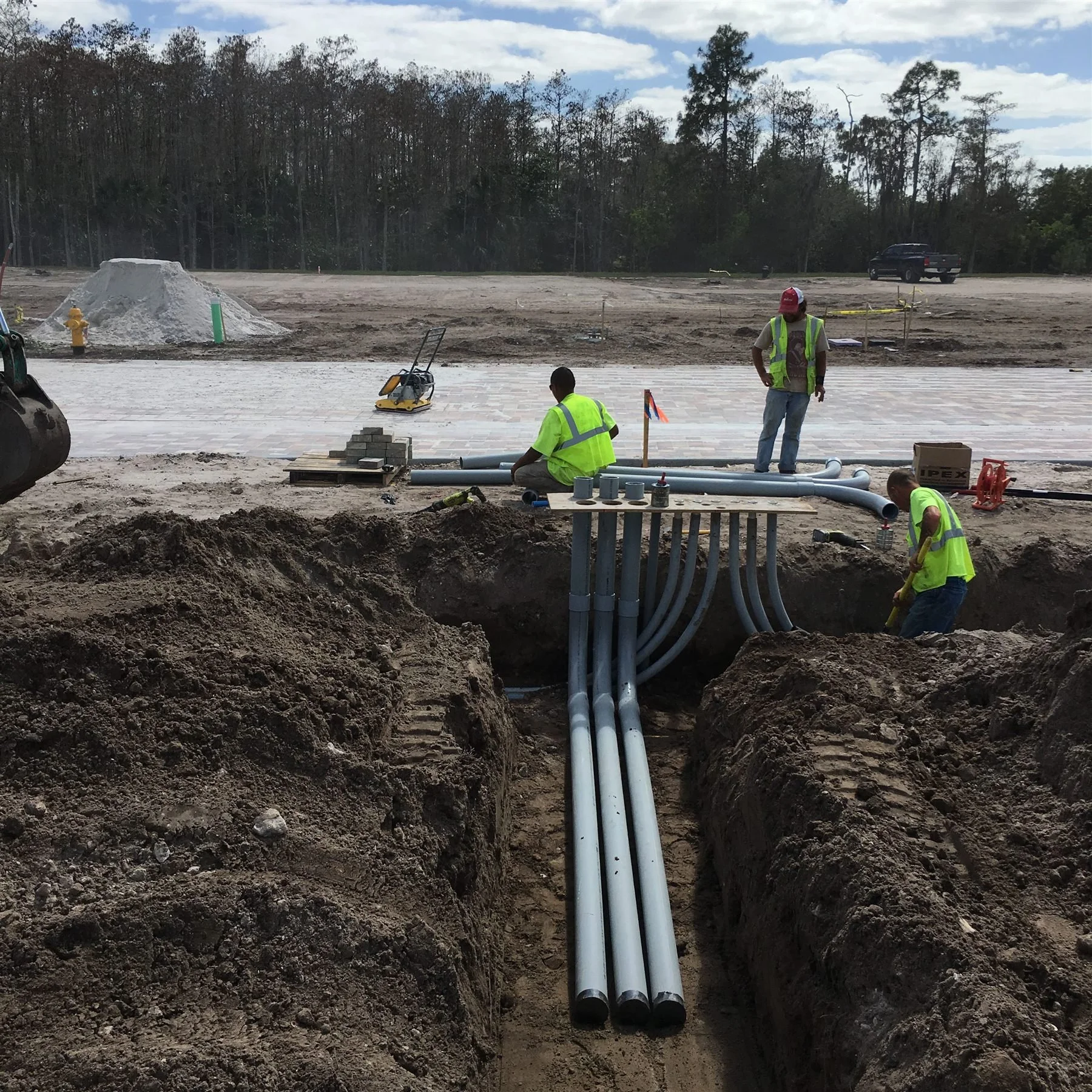 Construction workers installing underground piping on a construction site with trees in the background.