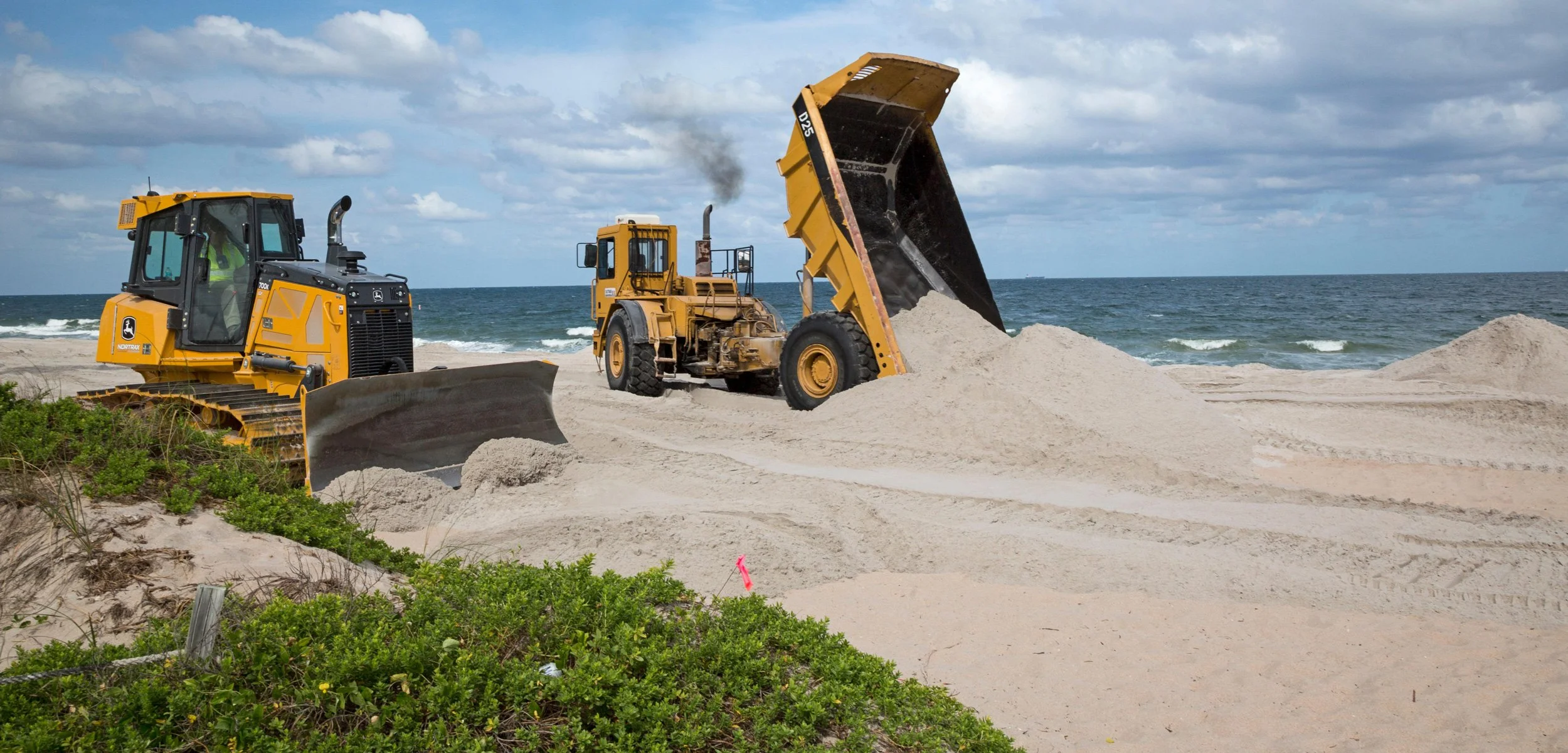 Construction equipment working on a beach, with a bulldozer and a front loader moving sand, near the ocean under a cloudy sky.