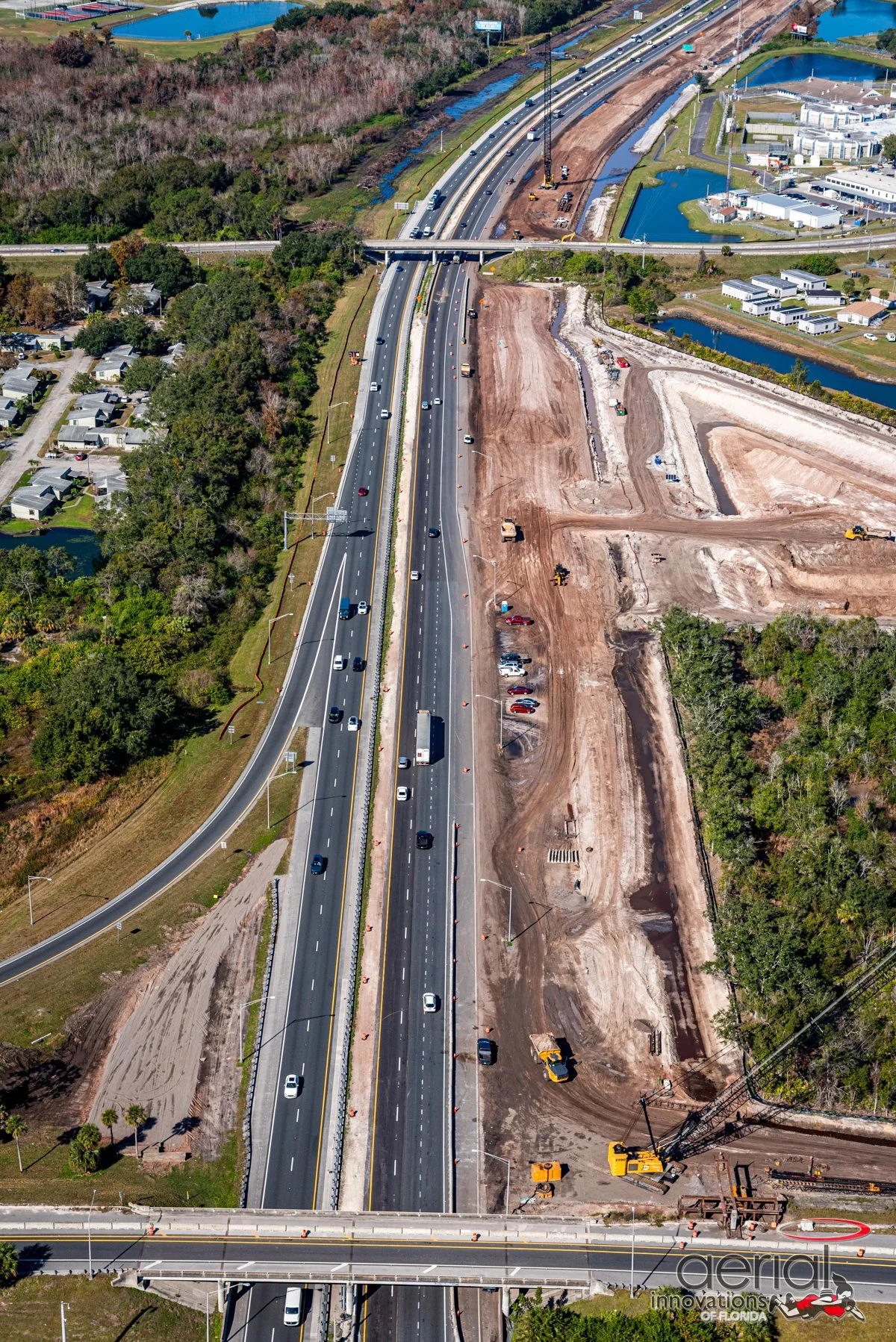 Aerial view of a road construction site with multiple lanes, some lanes open with vehicles, some under construction with dirt and equipment. There is a bridge crossing over the main road, a mix of greenery and residential or commercial buildings near