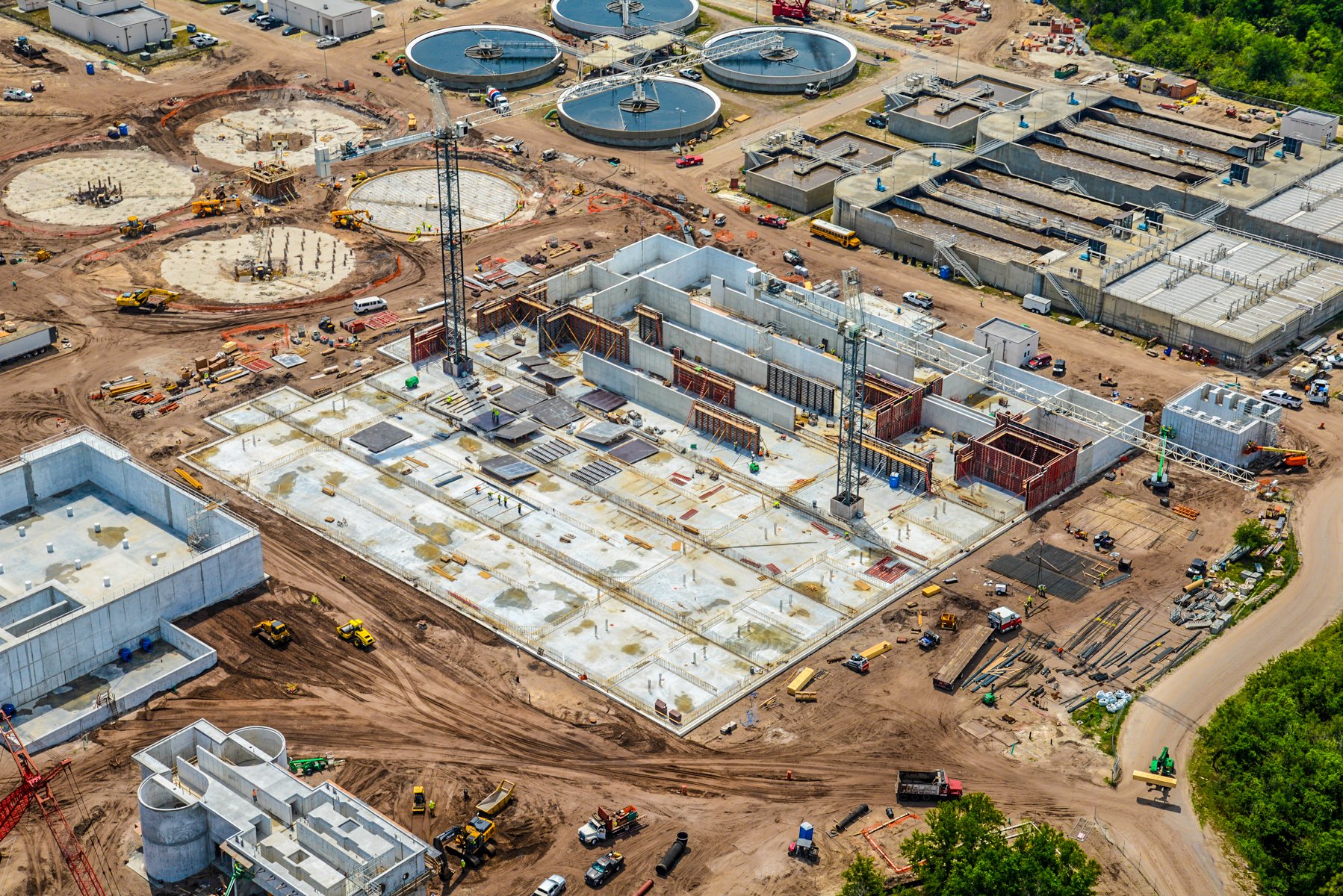 Aerial view of a large construction site with multiple structures in various stages of development, including building foundations, partially constructed walls, water treatment tanks, cranes, construction vehicles, and dirt roads.