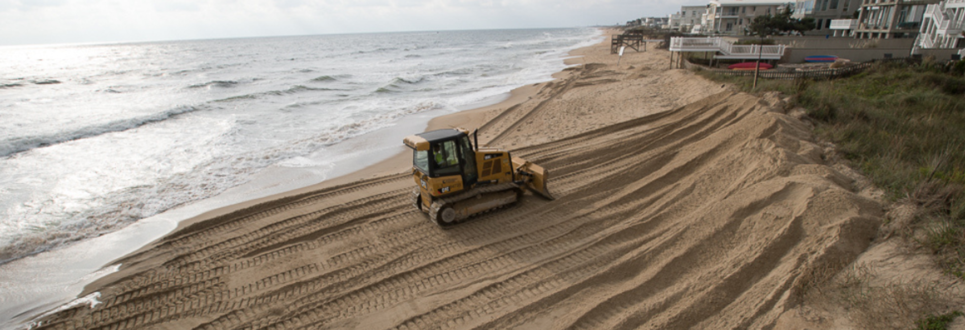 A bulldozer smoothing sand on a beach near ocean waves with condominium buildings in the background.
