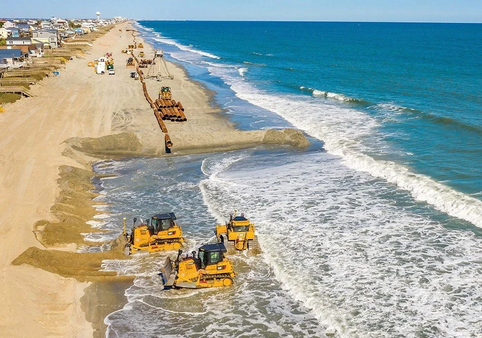 Construction equipment, including bulldozers and pipes, working on a beach to rebuild or repair the shoreline.
