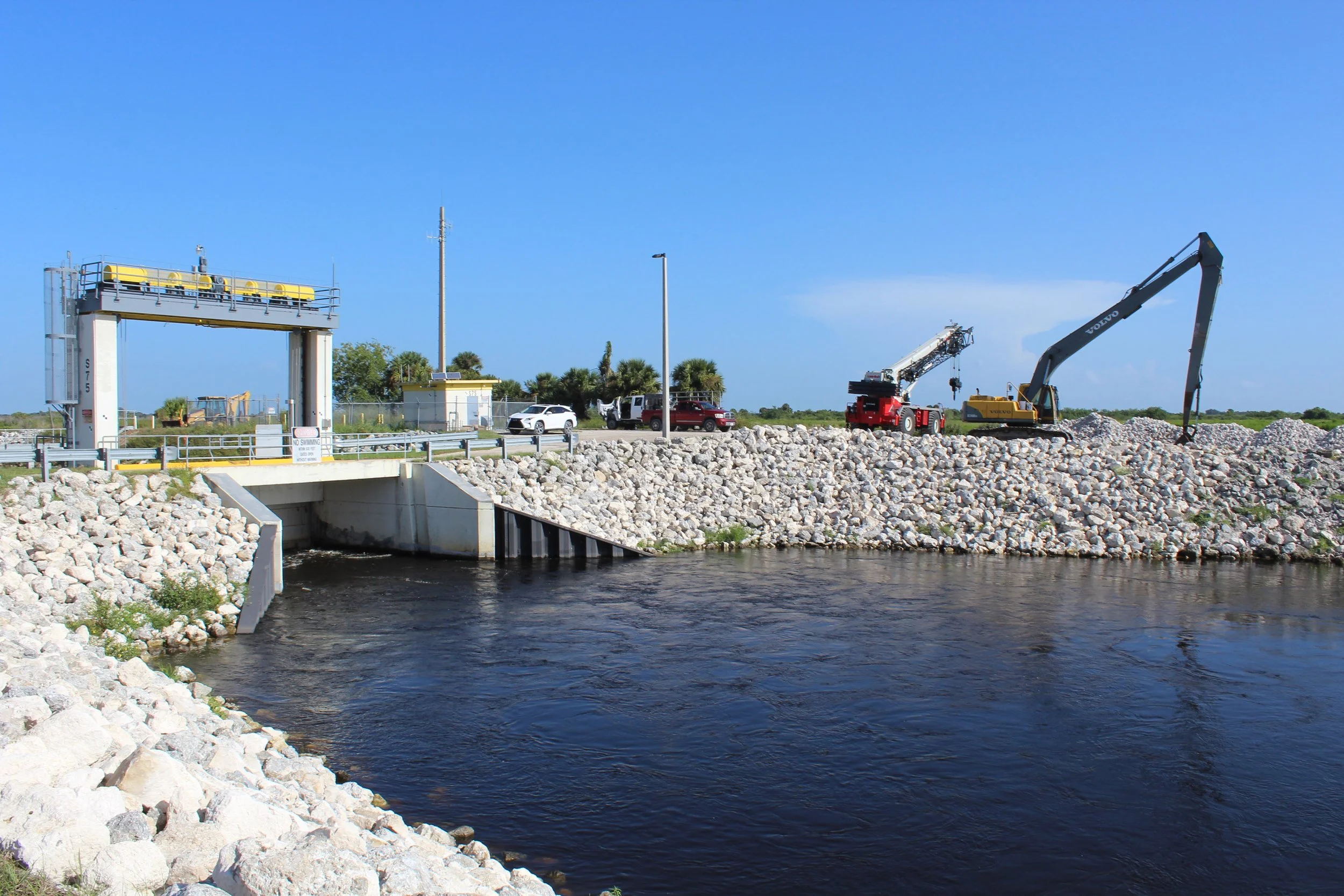 View of a dam area with a body of water over which a control gate is positioned, and two construction cranes nearby, with vehicles parked on the road and a small building, under a clear blue sky.