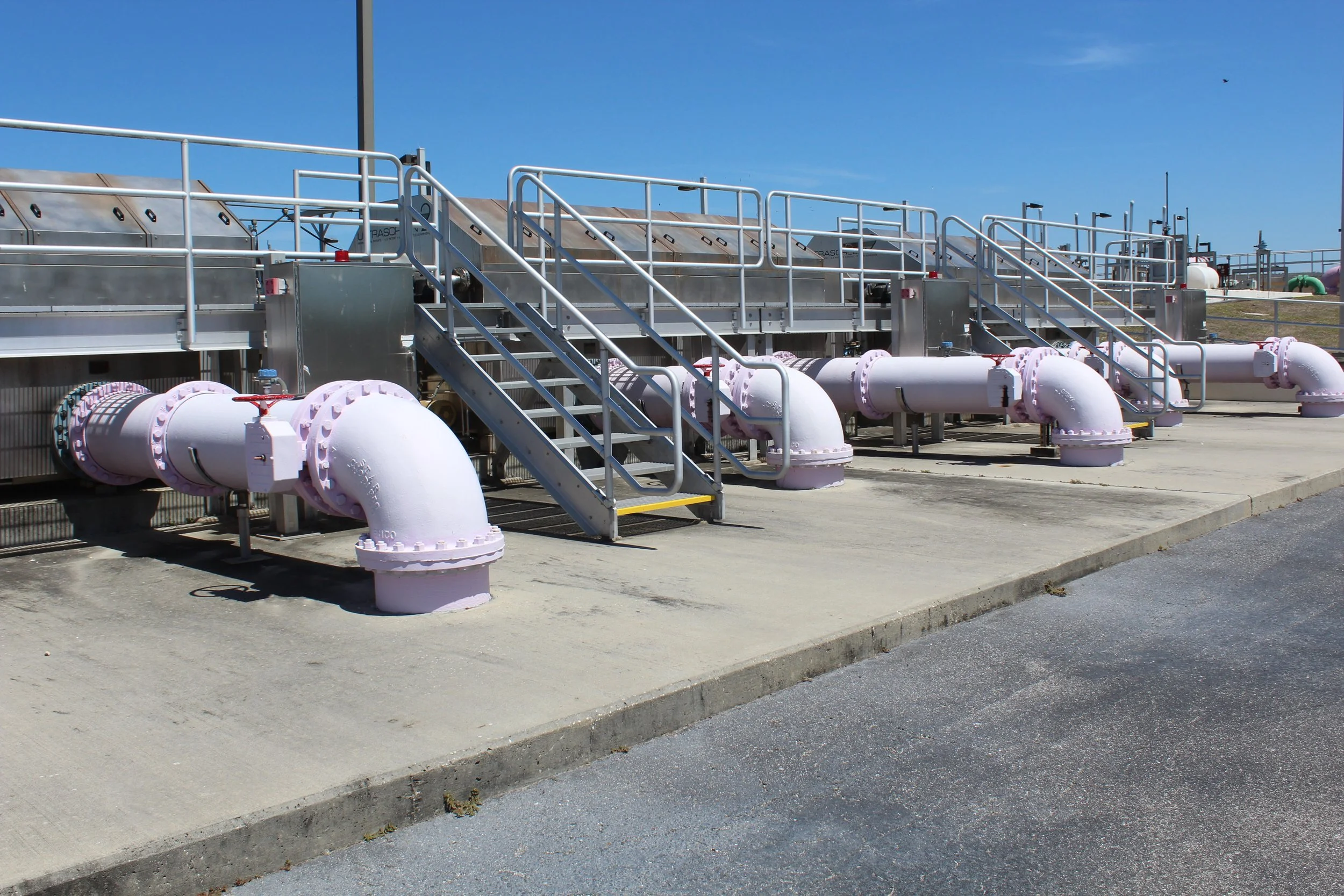 Industrial facility with metal stairs and large white pipes installed on a concrete ground, under a clear blue sky.