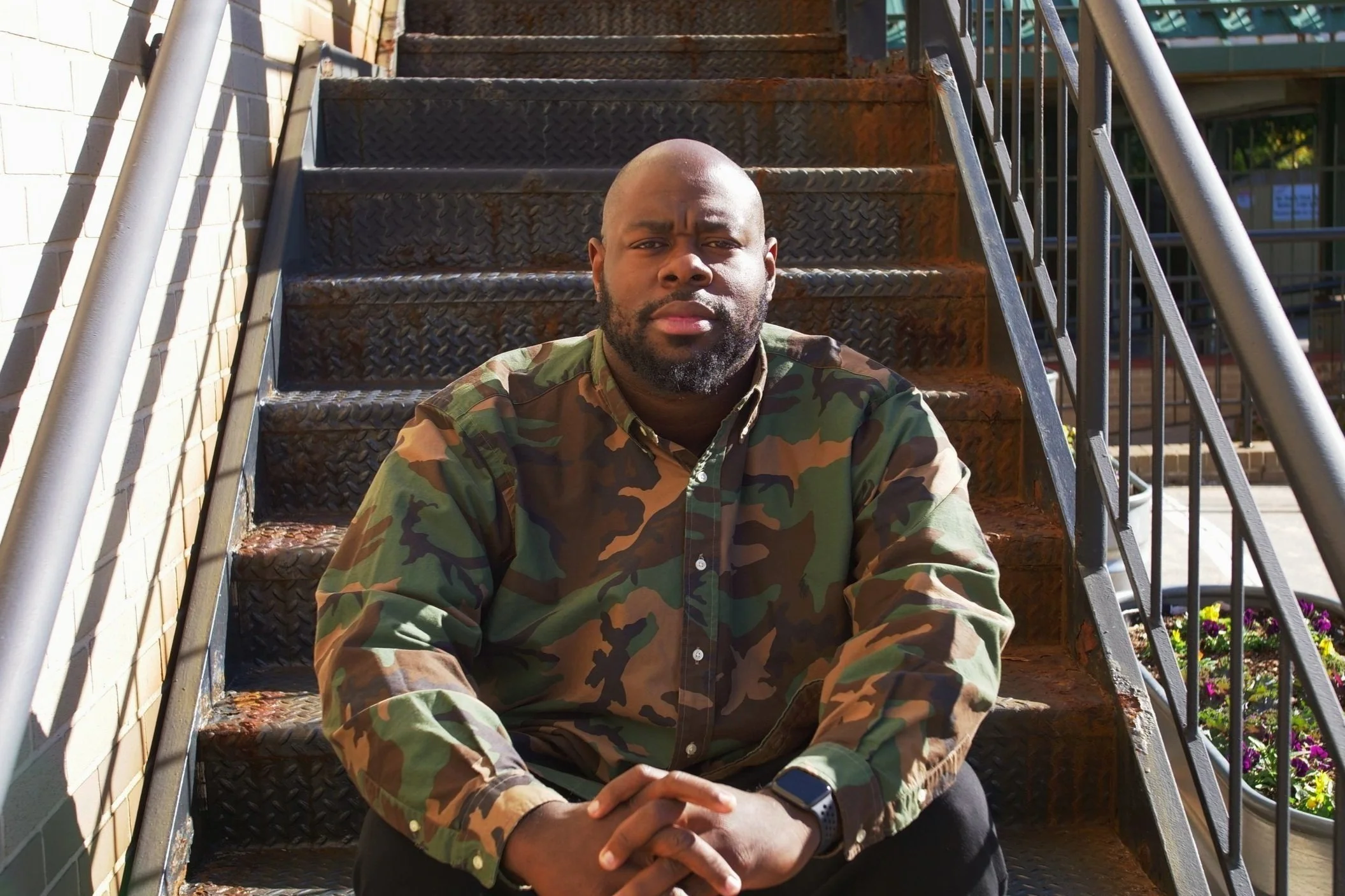 Charlie Mitchell sitting on outdoor metal stairs, wearing a camouflage shirt and a wristwatch, with a serious expression, in sunlight.