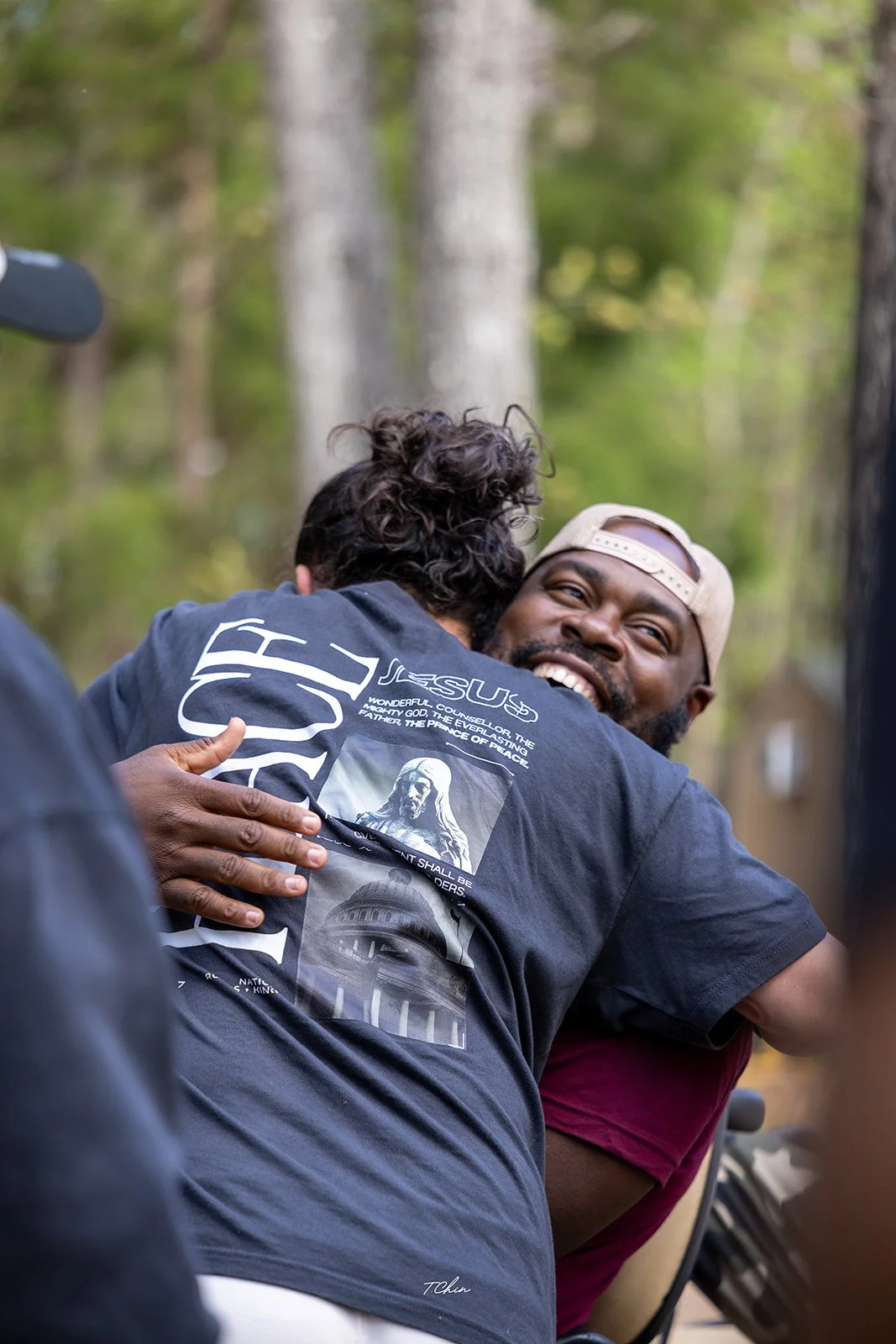 Two men hugging outdoors, one smiling widely and wearing a beige cap backwards.