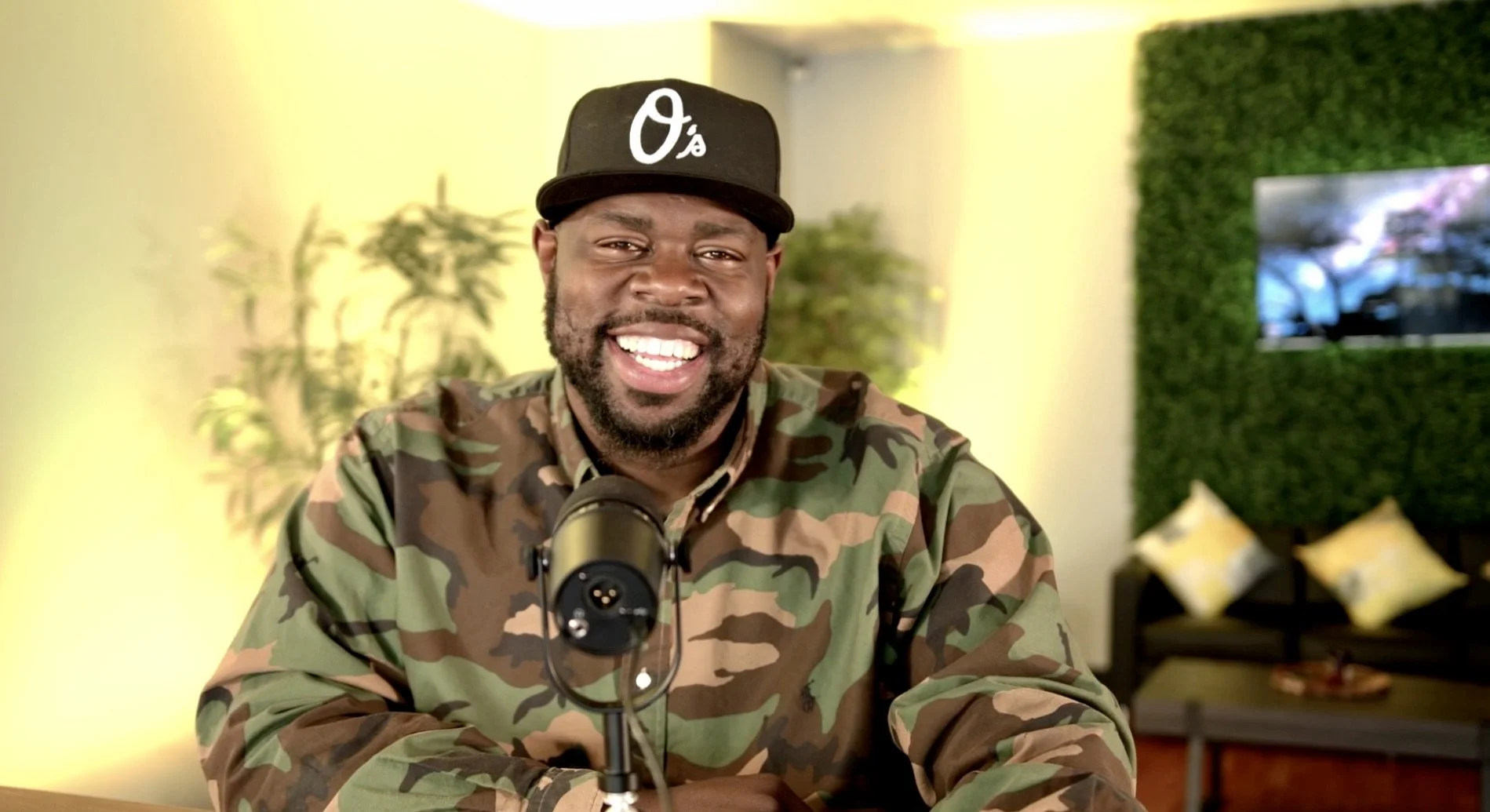A smiling man with a beard wearing a camouflage shirt and a black baseball cap with a white A's logo, sitting at a table with a microphone in front of him in a brightly lit room with green plants and a TV on a wall in the background.